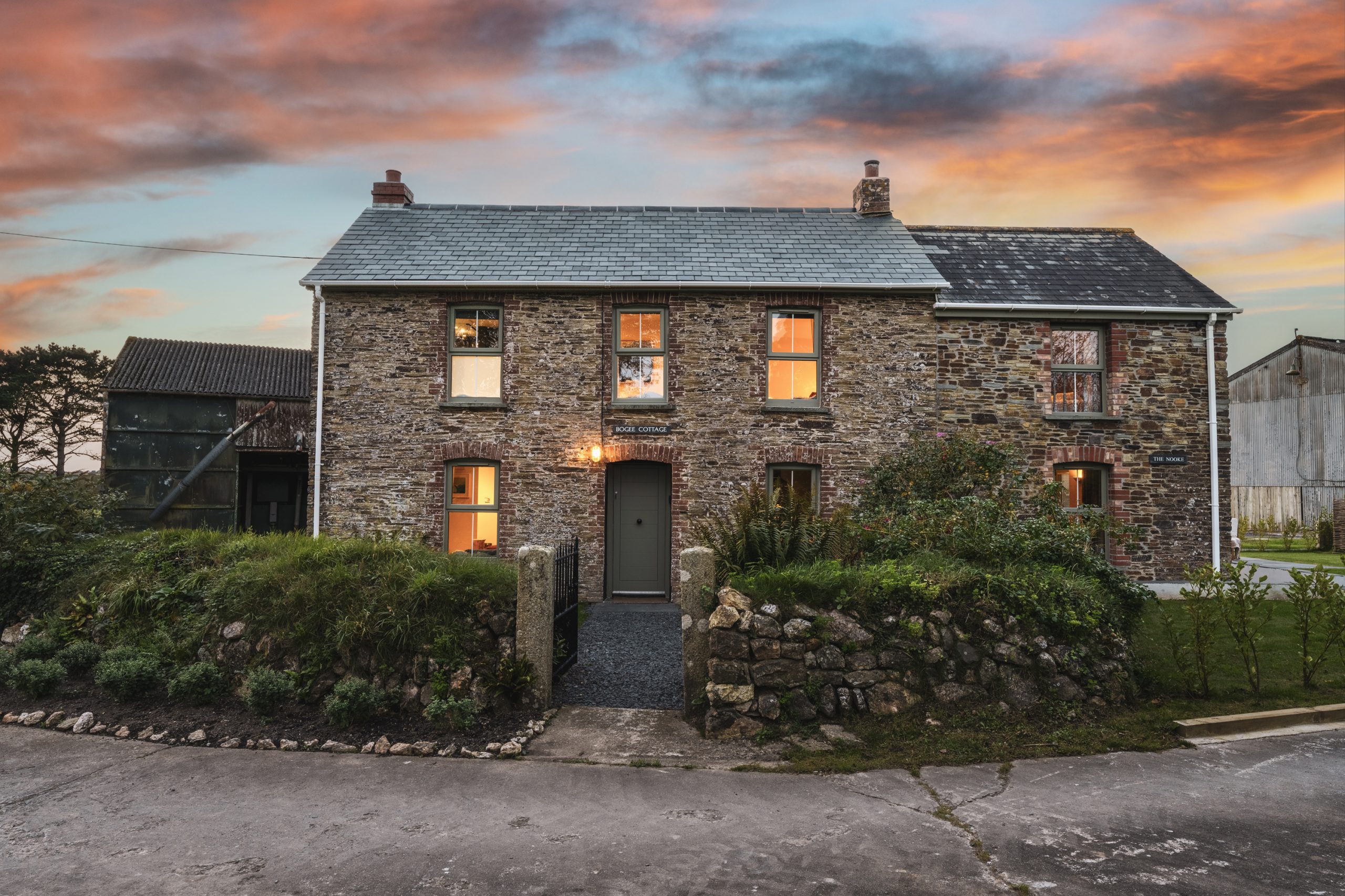 A cheap romantic escape awaits in a stone farmhouse with lit windows at sunset, surrounded by lush greenery and a rustic stone wall, while a charming barn stands nearby.