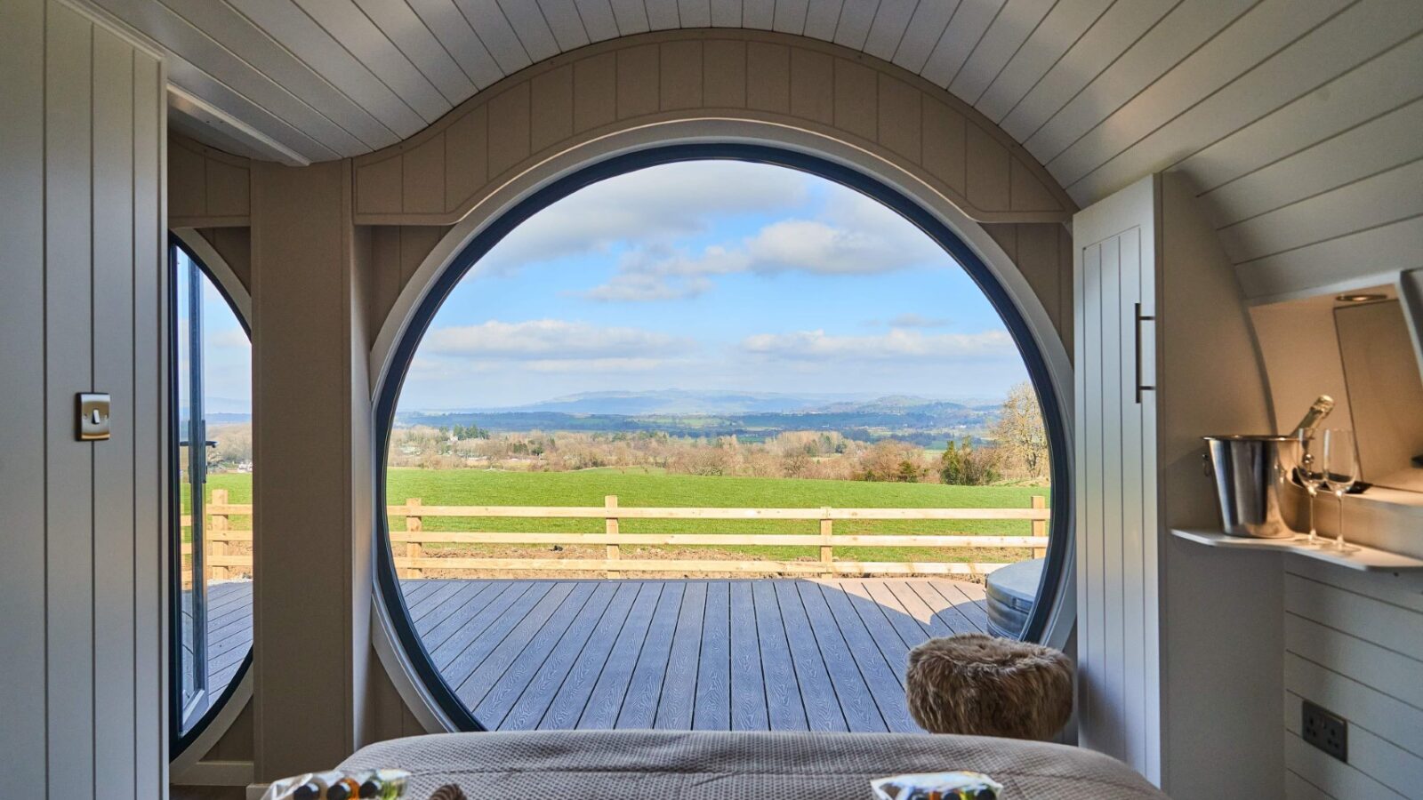 The circular window offers a breathtaking view from the cozy cabin interior, overlooking a wooden deck and grassy field stretching toward the distant hills of Camlad Valley beneath a cloudy sky.