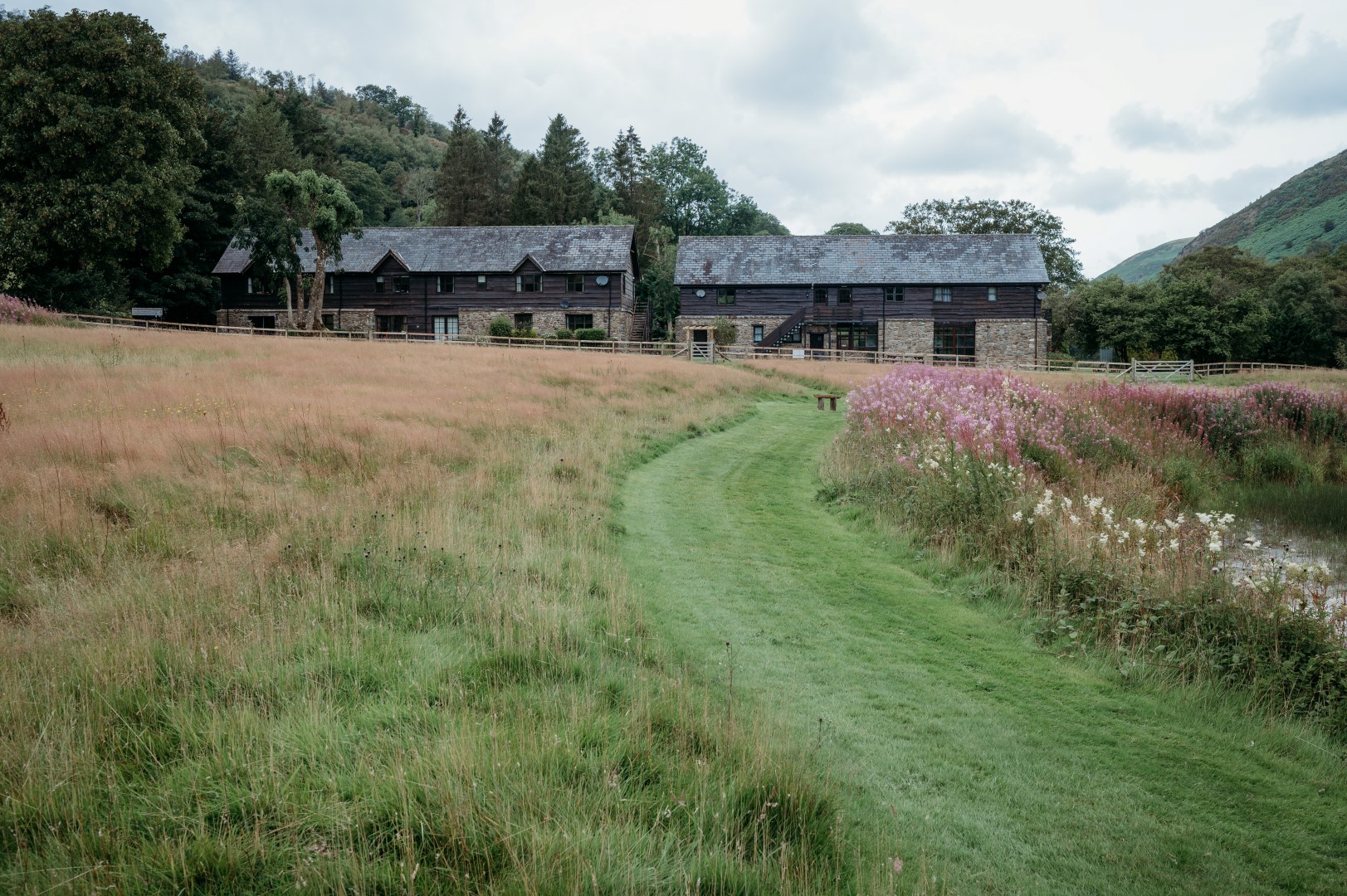 A path leads through a meadow to a row of rustic wooden buildings, offering a cheap romantic escape surrounded by trees and hills under a cloudy sky.