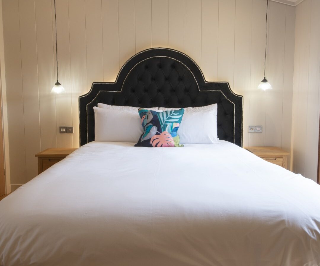 A neatly made bed with a black tufted headboard, white bedding, a colorful pillow, and two hanging bedside lamps offers a cozy feel reminiscent of serene lakeside lodges.