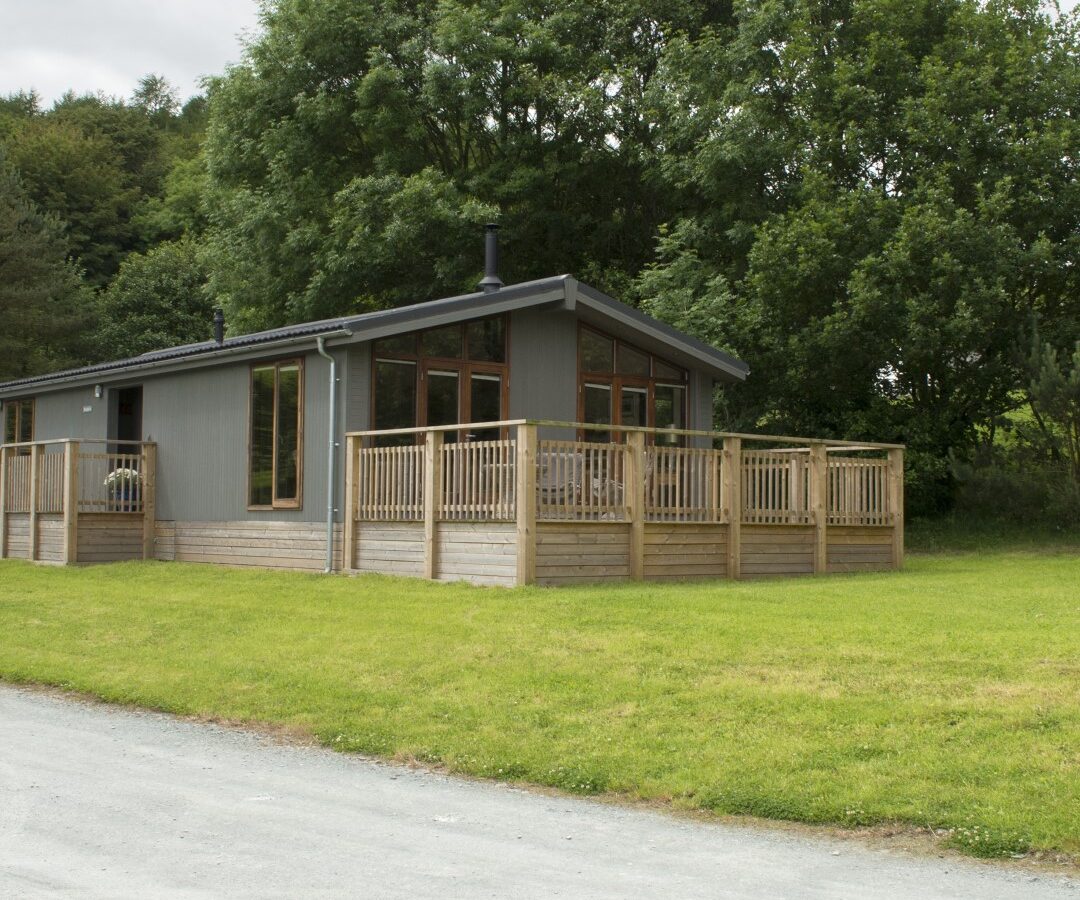 A modern lodge with large windows and a wooden deck, surrounded by grass and trees on a cloudy day, offers serene views that make it feel like you're nestled beside a tranquil lake.