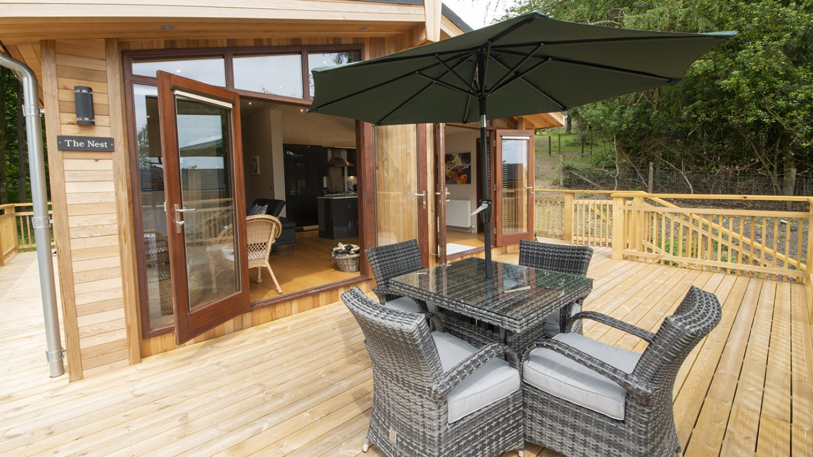 Wooden deck with wicker dining set and green umbrella, perfect for lakeside lodges. Open double doors lead to greenery-filled interior.