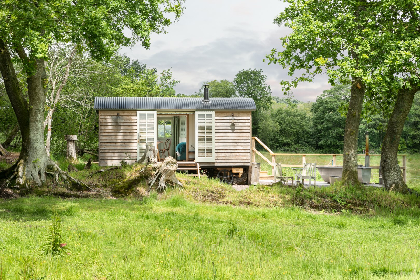 A small wooden cabin with open double doors, nestled among trees and grass, offers a perfect retreat for cheap romantic holidays. A charming deck on the right side invites you to unwind in nature's embrace.