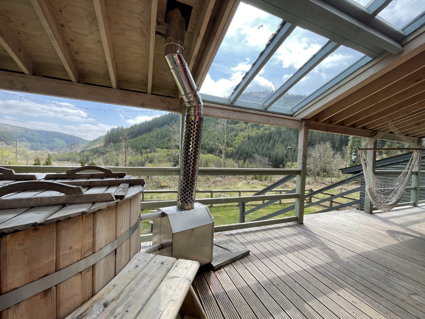 Enjoy a cheap romantic getaway on a wooden deck with a hot tub and hammock chair, offering enchanting views of green hills and a cloudy sky through a slanted glass roof.