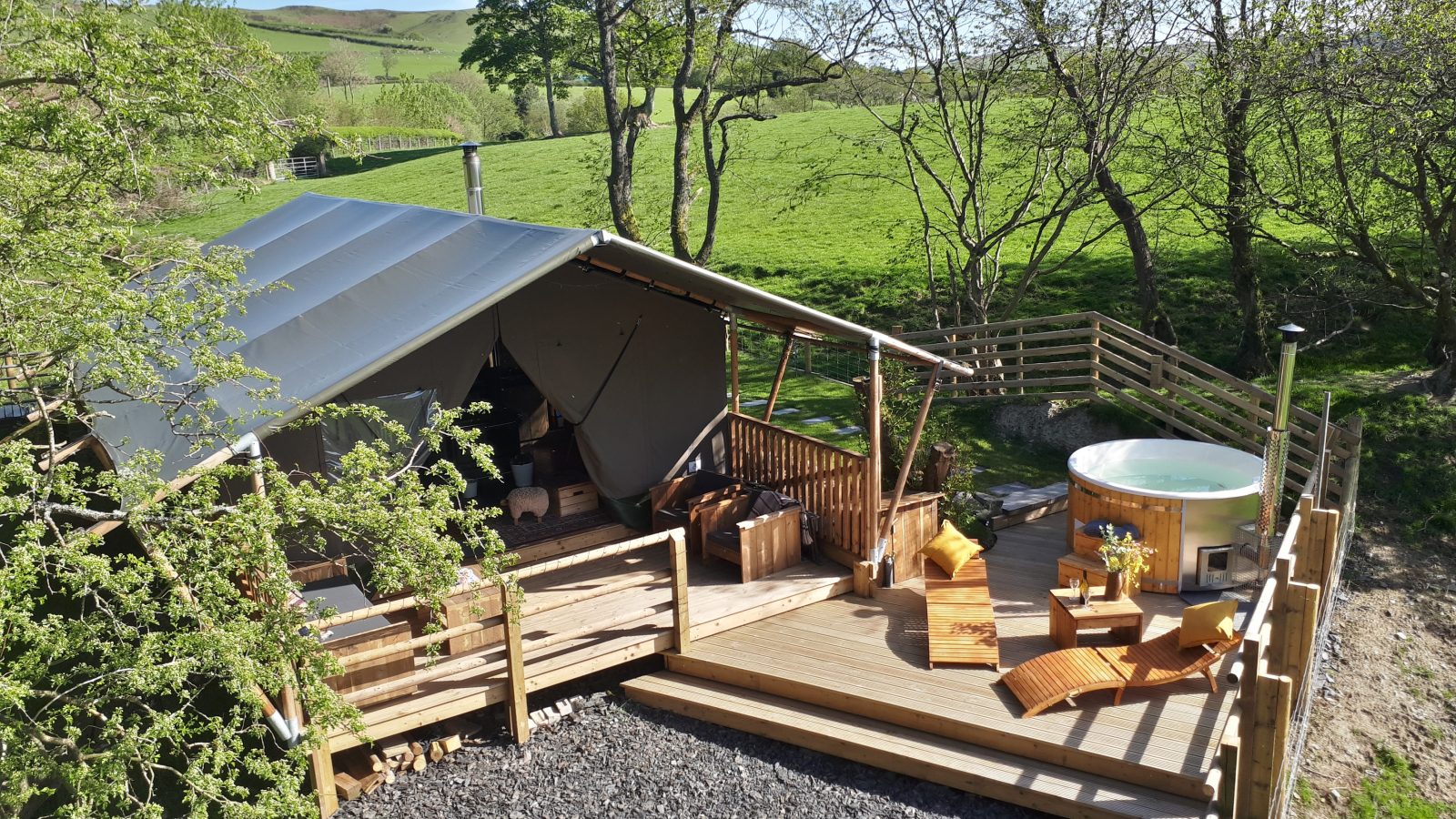 A glamping tent with a wooden deck, lounge chairs, and a hot tub awaits you amidst the scenic countryside of Wales, surrounded by lush green fields.