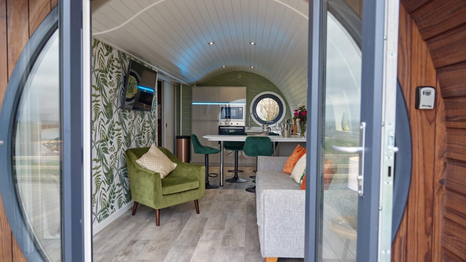 Experience the Camlad Valley Luxury Pods, where the interior of a small, modern living space invites you in through its round entrance. Inside, a green chair and gray sofa complement the chic kitchen area, offering an exquisite blend of comfort and style amidst serene surroundings.