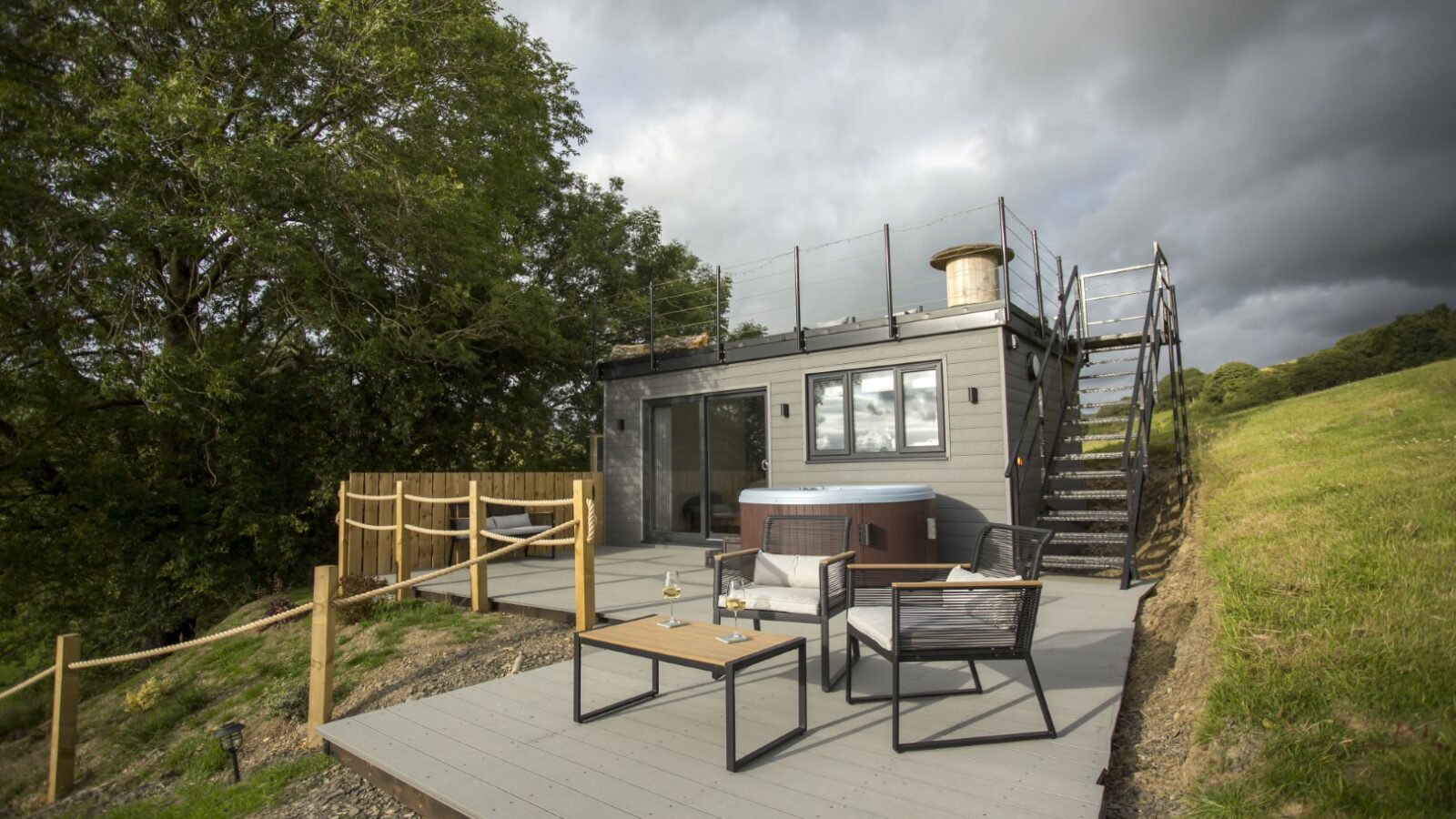 Cae Cnwc's charming rooftop cabin offers a patio, hot tub, and cozy seating area atop the hillside under cloudy skies.