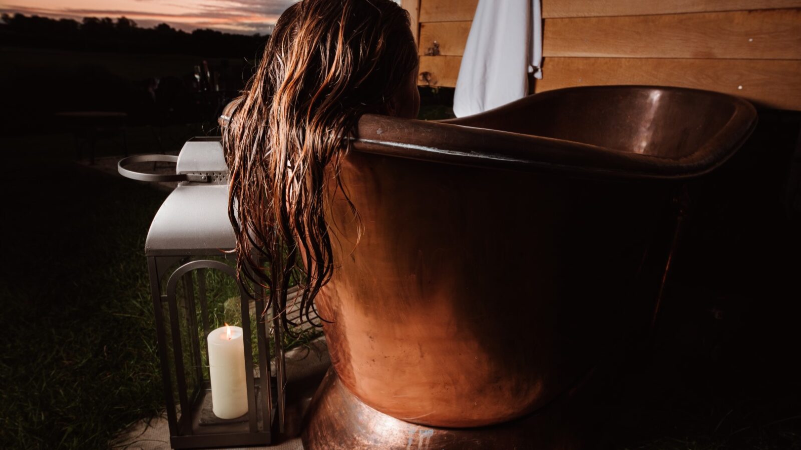 A person with wet hair relaxes in a bronze bathtub beside a lit candle lantern, enjoying the breathtaking sunset view—a perfect evening of Ewe Glamping.