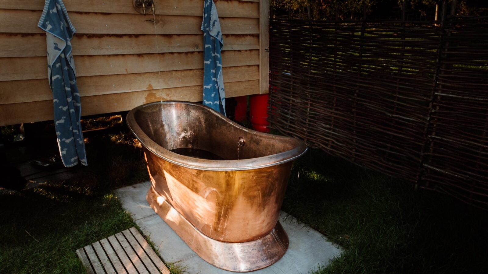 A copper bathtub is placed outdoors on a concrete slab with two blue towels hanging on a wooden wall nearby, creating an ideal Ewe Glamping experience.