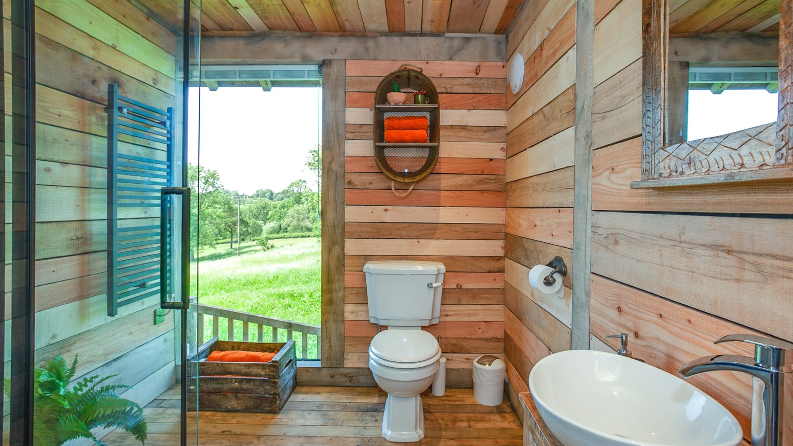 The Cadwollen Treehouse bathroom exudes rustic charm, featuring a wooden design with a toilet, white sink, and mirror. Towels rest neatly on shelves, while the glass shower enclosure and window offer scenic views of green fields. A leafy plant adds a touch of greenery to this serene space.