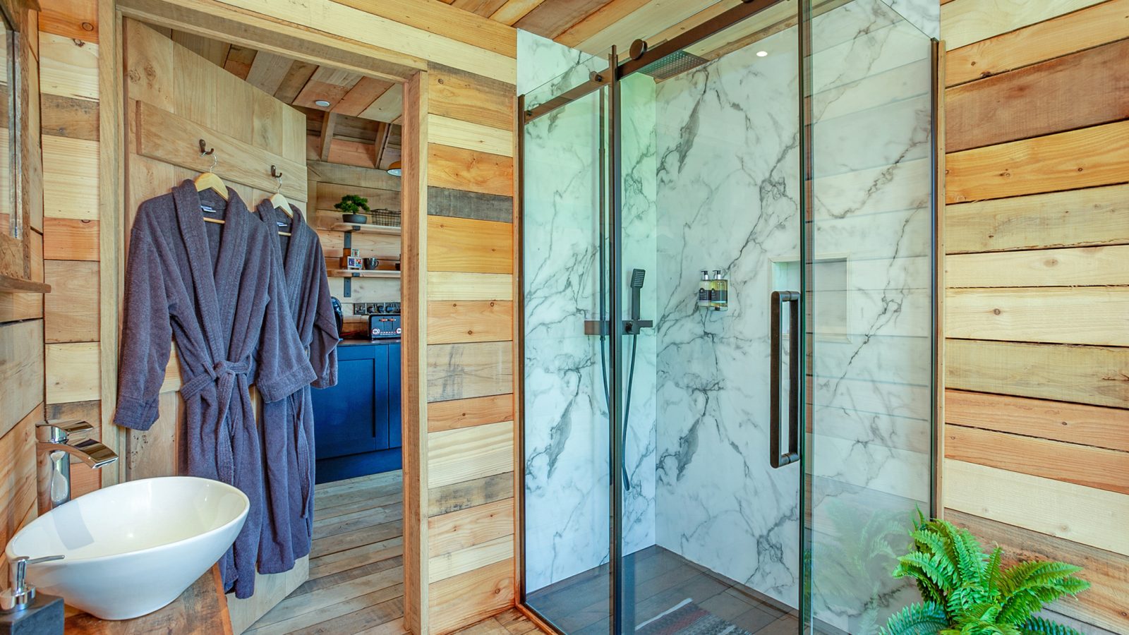 The Cadwollen Treehouse offers a unique accommodation experience with a modern bathroom featuring wood-paneled walls, a glass shower with marble tiles, and a round sink on a wooden counter. Two dark bathrobes hang on the door, while a green plant adds a touch of nature to this enchanting treehouse rental.