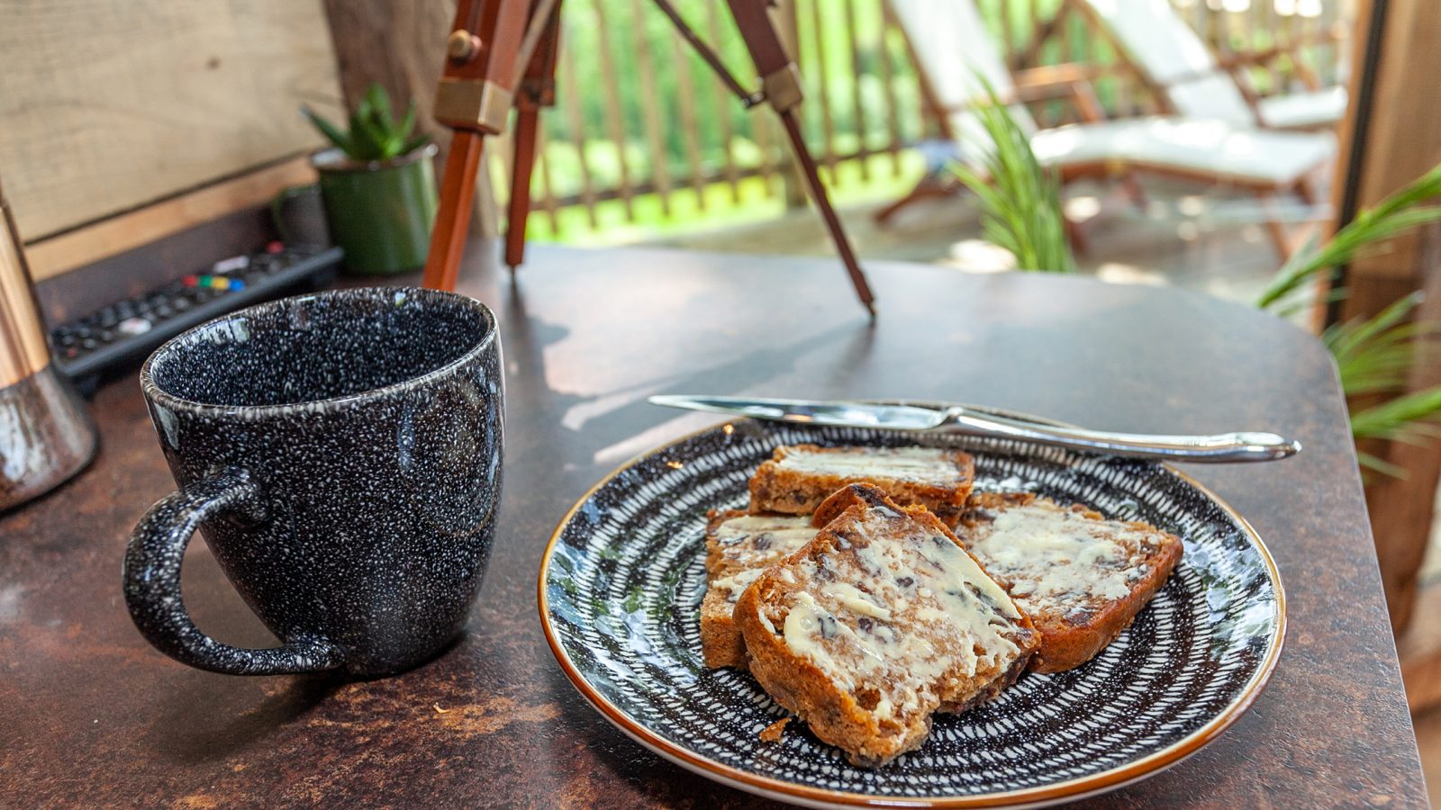 A black speckled mug sits on a brown table beside a patterned plate with slices of buttered bread, all under the rustic charm of the Cadwollen Treehouse. A knife rests faithfully by the plate while outdoor chairs and a tripod peek through the railing in the background.