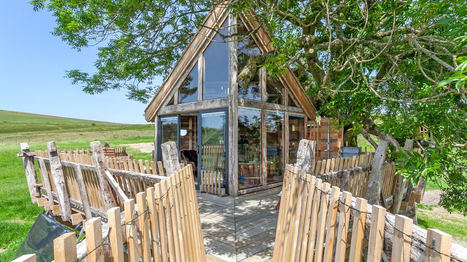 The Cadwollen Treehouse is a charming wooden retreat with large glass windows nestled among green foliage. Featuring a pointed roof, it’s accessed by a wooden walkway with railings. This accommodation sits in a grassy, open area under a clear blue sky, offering tranquility and nature's embrace.