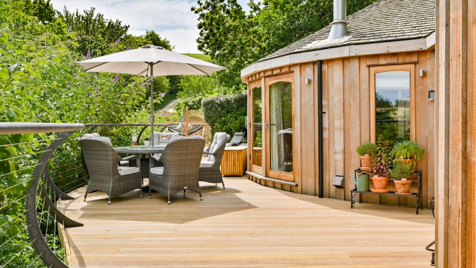 A charming wooden deck featuring wicker chairs and a table under an umbrella sits next to a round wooden building surrounded by lush greenery, reminiscent of the serene Dittisham Hideaway treehouses.