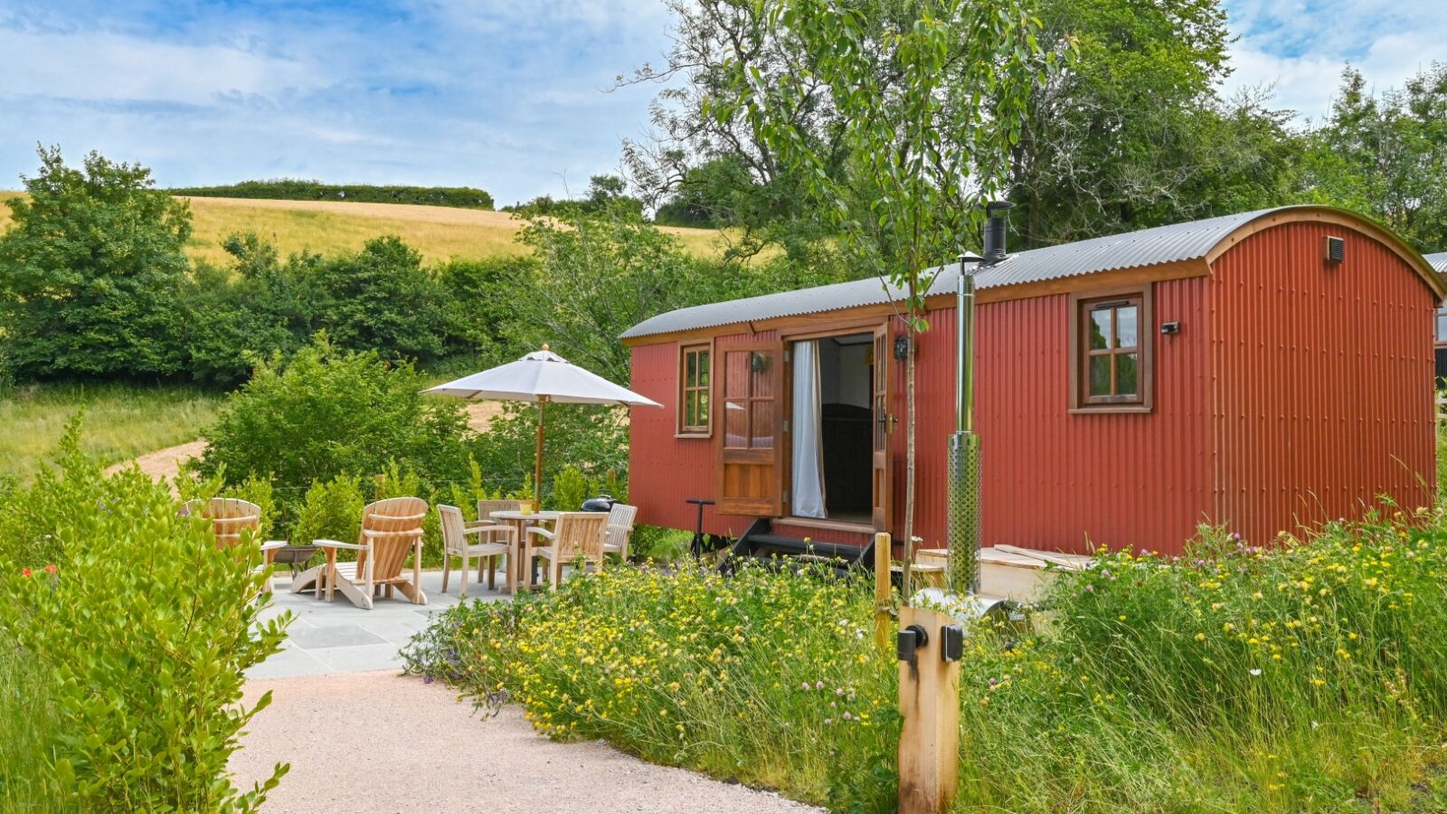 A charming red cabin at Dittisham Hideaway nestles in a verdant landscape, featuring outdoor seating and an umbrella along a gravel path. This picturesque setting under a blue sky with scattered clouds mirrors the rustic allure of traditional Shepherds Lodges.