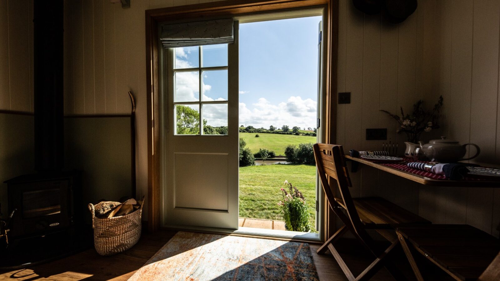 The view from inside a serene glamping accommodation features an open door leading to a sunny field, complete with a cozy table, chairs, and a basket nestled by the stove.