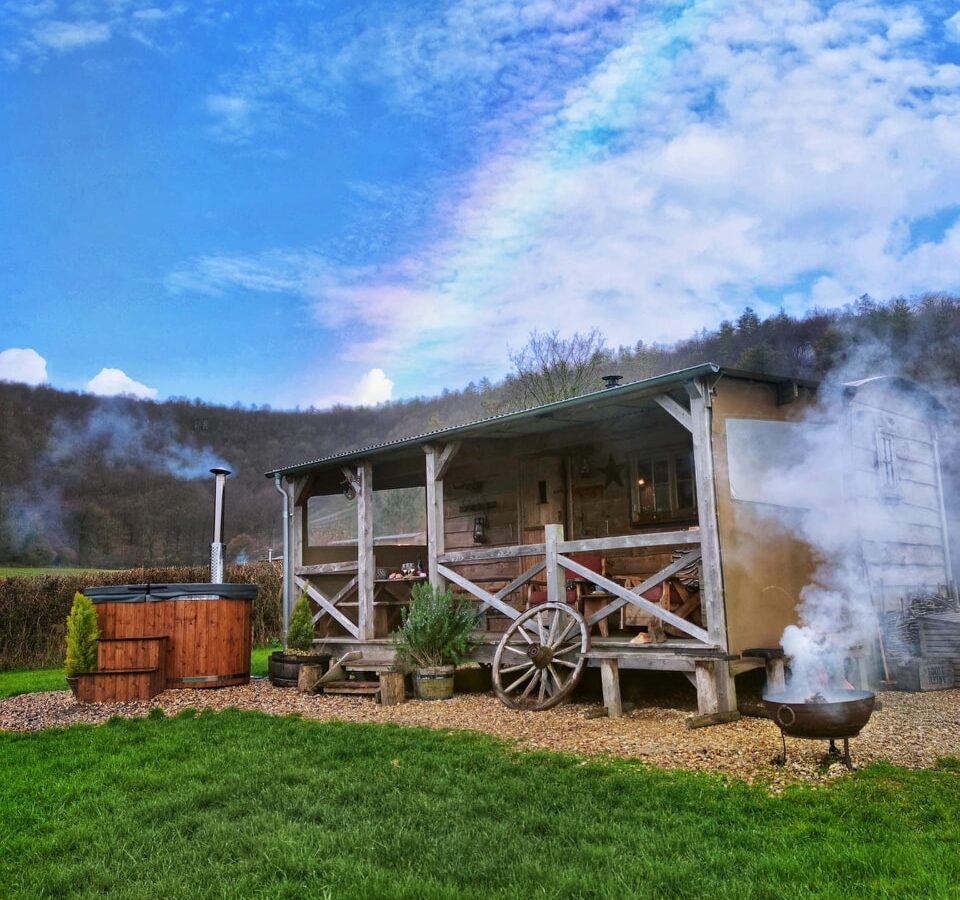 A rustic cabin with a porch, nestled among grass and trees, evokes the charm of a Loose Reins ranch. In the yard, a steaming hot tub and fire pit invite relaxation and warmth under the open sky.