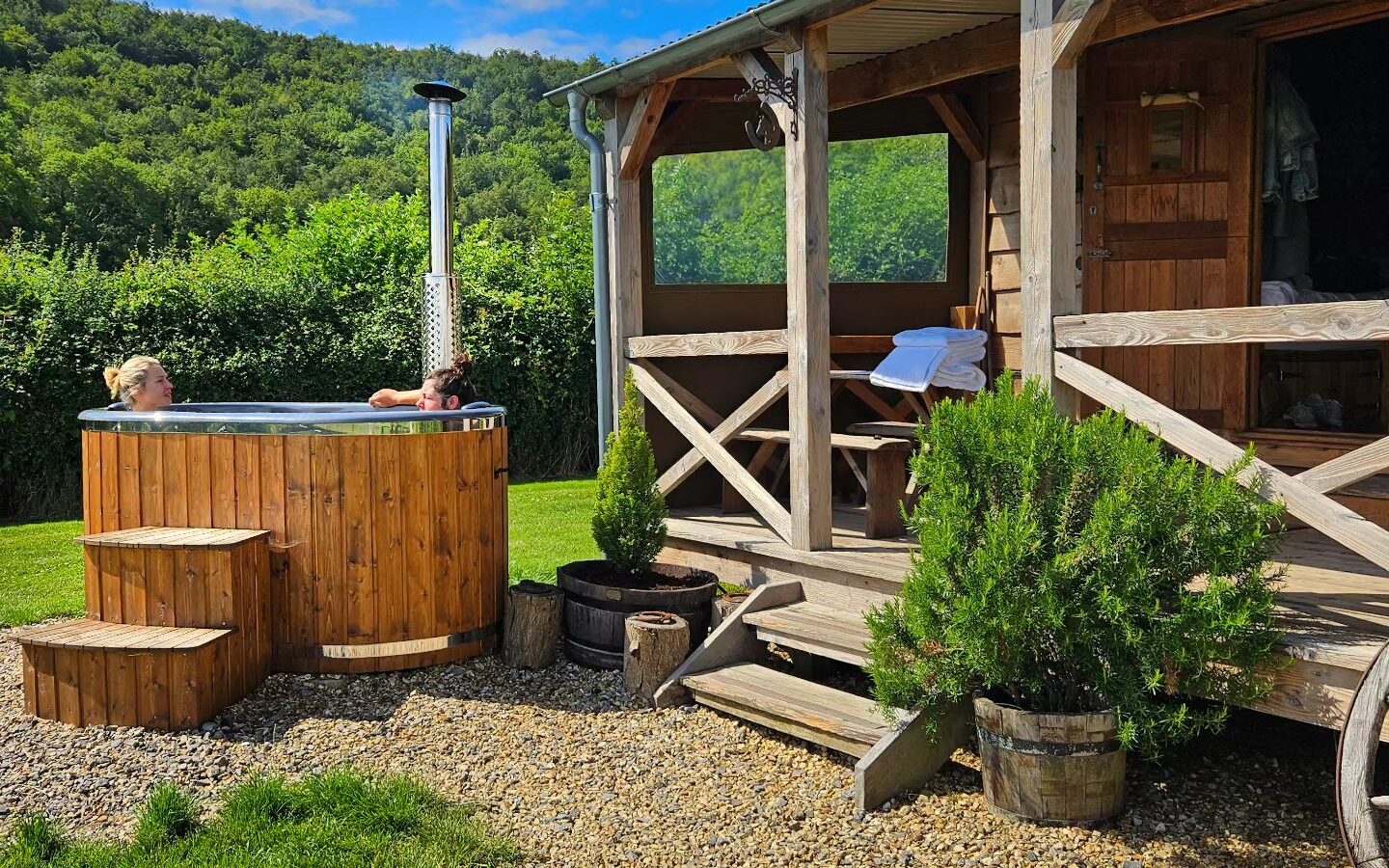 A woman unwinds in a wooden hot tub beside a charming cabin at Loose Reins Ranch, surrounded by lush greenery and a clear blue sky.