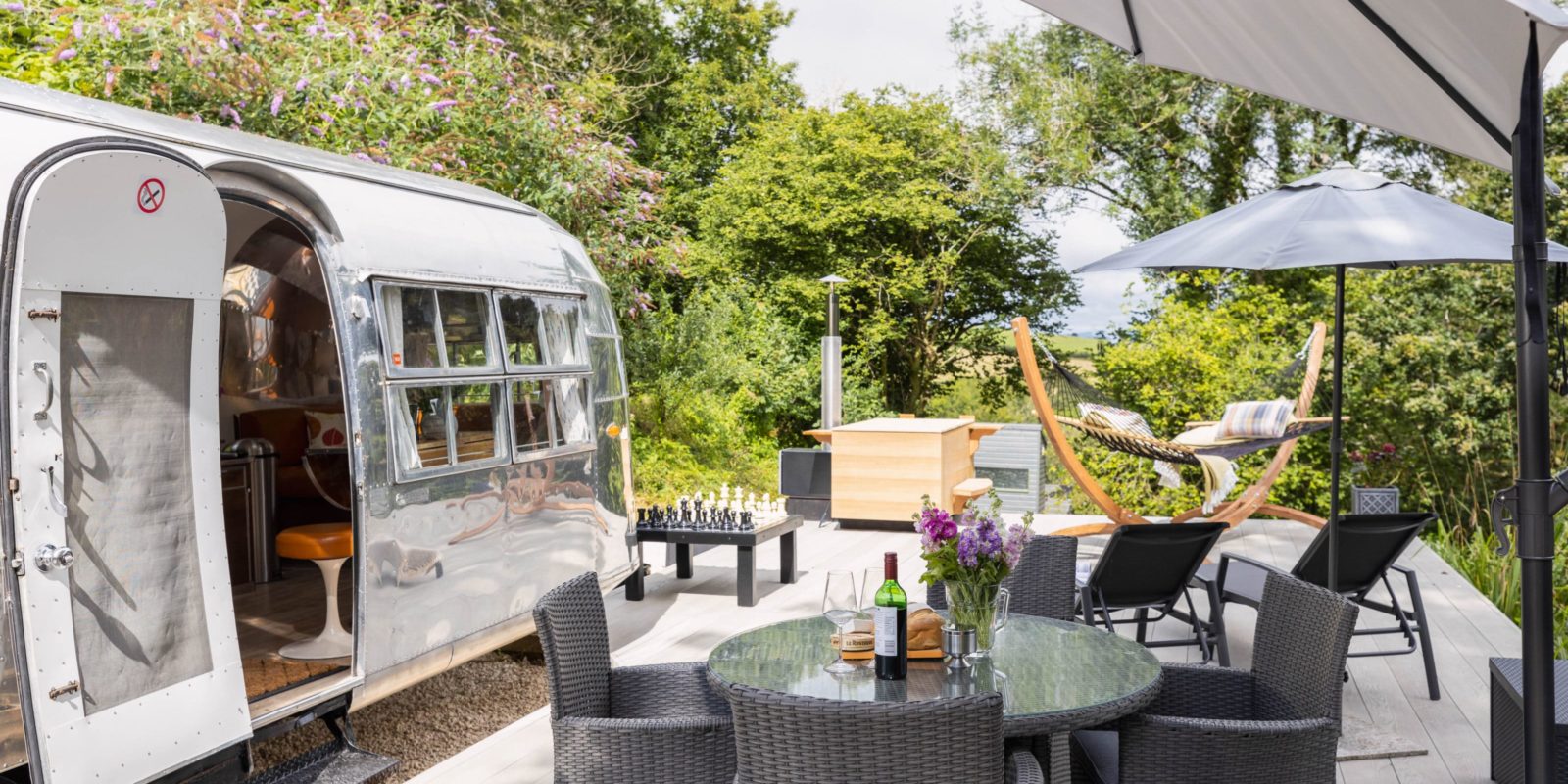 An outdoor patio in Dittisham features an American Airstream, round table and chairs, hammock, umbrella, chessboard, and lush greenery in the background.