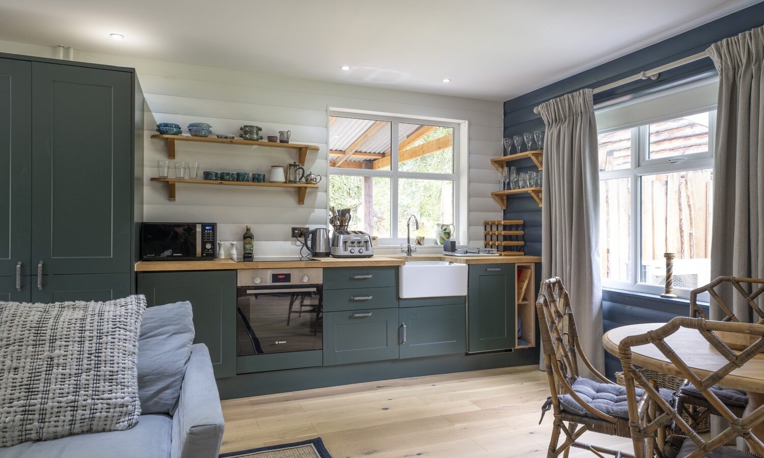 A cozy kitchen with green cabinets, open shelves, a farmhouse sink, and a small dining area with wicker chairs by the window evokes the charming atmosphere of Keepers Bothy.