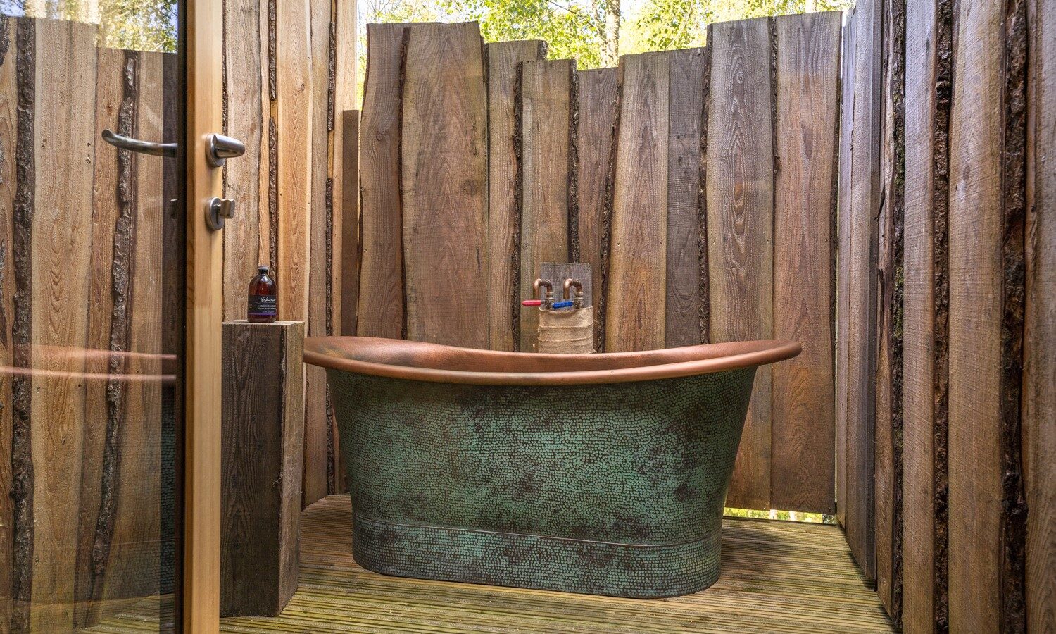 An outdoor bathroom reminiscent of a treehouse, featuring a green metal bathtub surrounded by wooden fencing with trees and flycatchers fluttering in the background.