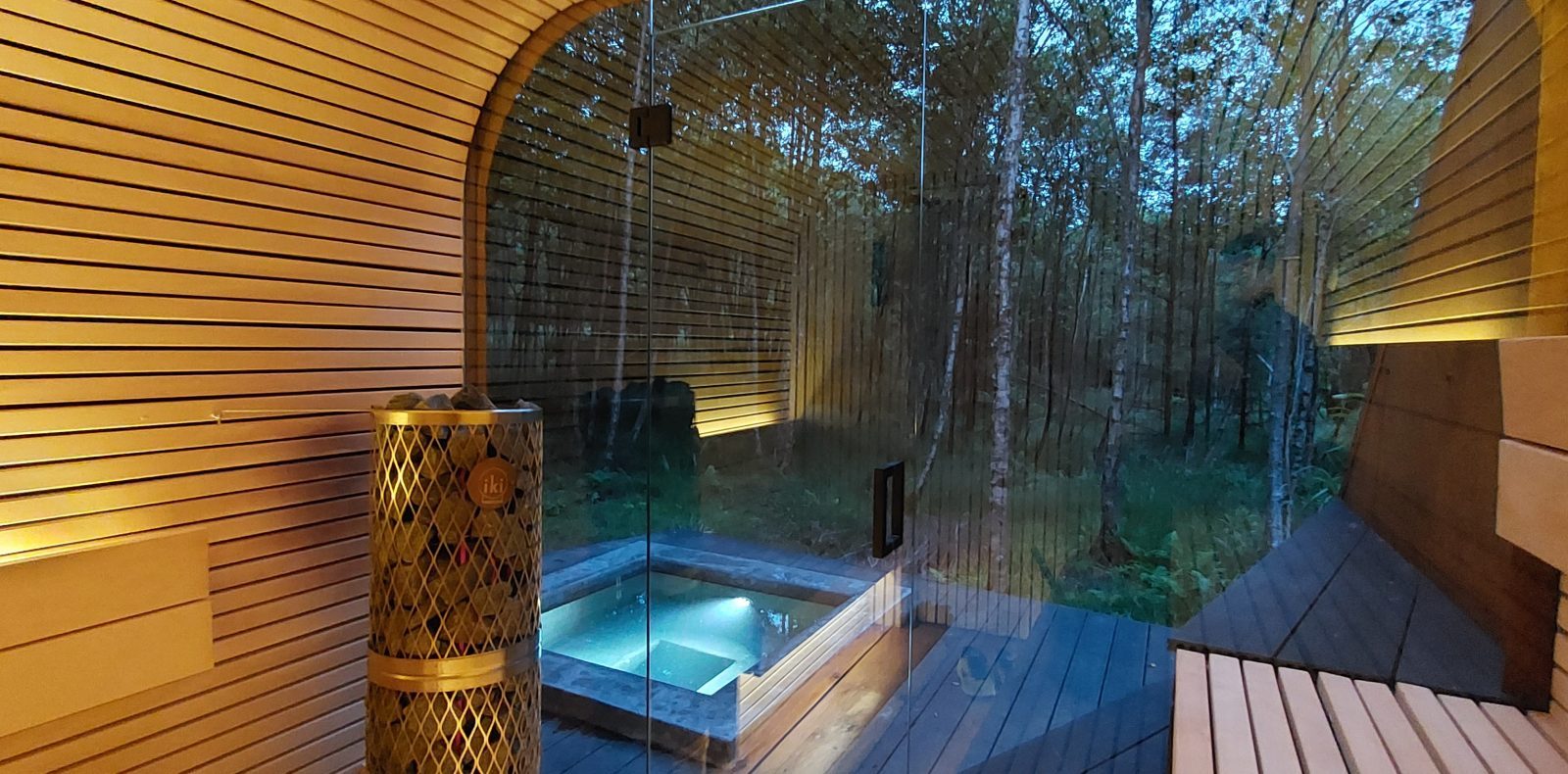 A modern treehouse sauna with wooden walls and a glass door overlooks a serene forest. Inside, there is subtle mood lighting and a small tiled hot tub on a wooden deck. The view outside, reminiscent of the Flycatcher Treehouse, reveals lush greenery and tall trees.