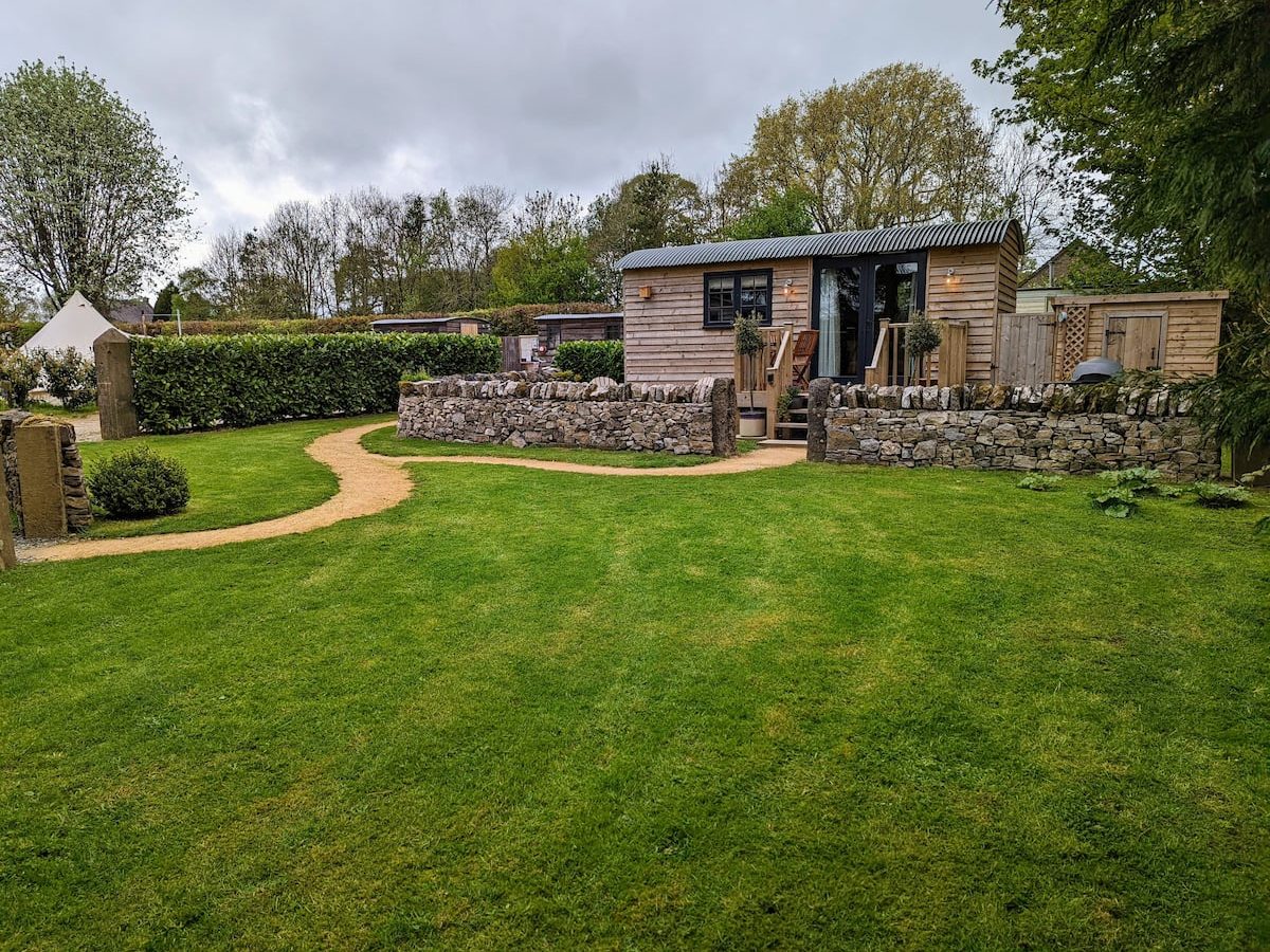 A well-maintained garden with a lush green lawn, a stone pathway, and a wooden cabin with a corrugated metal roof in the background. The scene is surrounded by hedges, trees, and cloudy skies reminiscent of the charm found in Smithyfields.