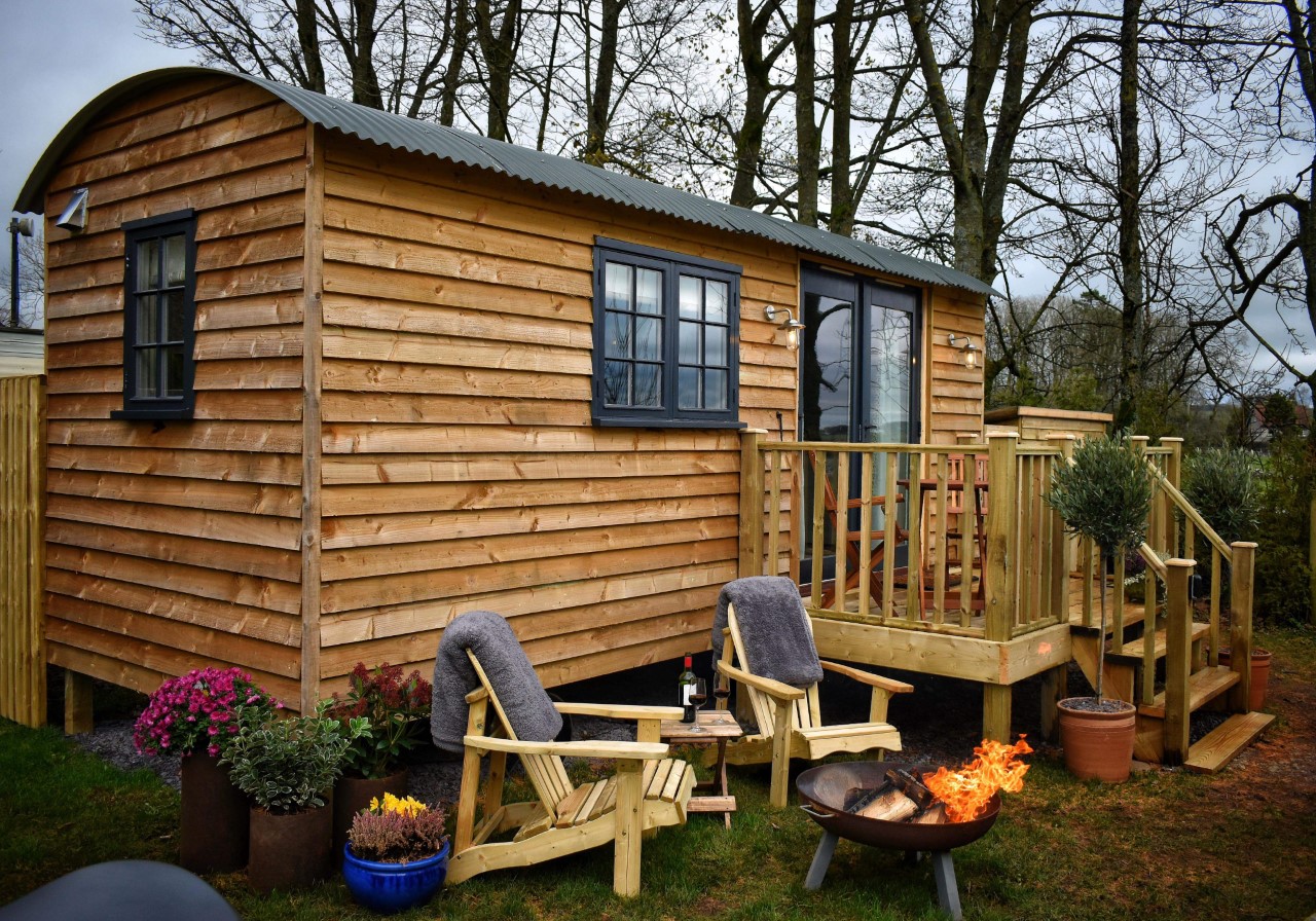 A cozy, small wooden cabin by Smithyfields with a corrugated metal roof. It features a deck with two wooden chairs, each draped with a grey blanket, a small table with a wine bottle and glass, potted plants, and a fire pit with a burning flame. The background has leafless trees reminiscent of rustic Shepherds Huts.