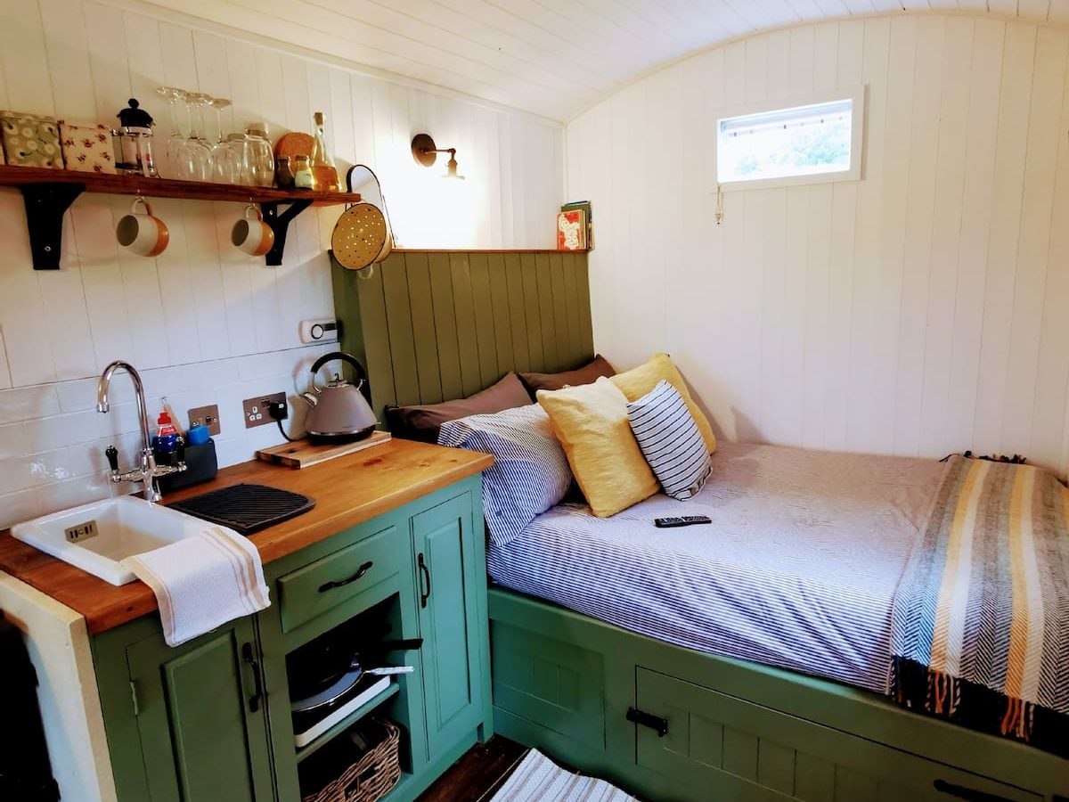 A cozy, compact bedroom and kitchen area with white walls and ceiling. The green kitchen in this Smithyfields Shepherds Hut features a counter, sink, and shelves with utensils and jars. The bed is snug with pillows and a blanket, and a small window above it allows natural light in. There is a rug on the wooden floor.