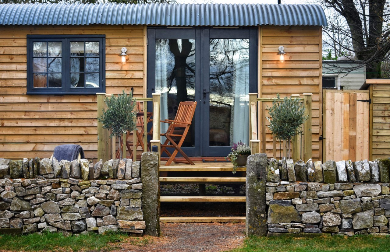 A cozy tiny house with wooden exterior, a small porch with two wooden chairs and a table, and a stone walkway leading to it. Stone walls with pillars frame the entrance. Reminiscent of Smithyfields Shepherds Huts, the house features large windows and is surrounded by trees and greenery.