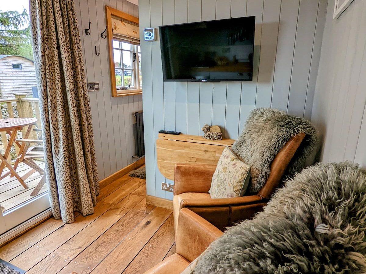 A cozy corner of a rustic room in the charming Smithyfields, featuring two brown leather armchairs with plush, furry throws. A small wooden table between the chairs holds a decorative stuffed animal. A flat screen TV is mounted on the gray-paneled wall, and a glass door opens to an outdoor patio.