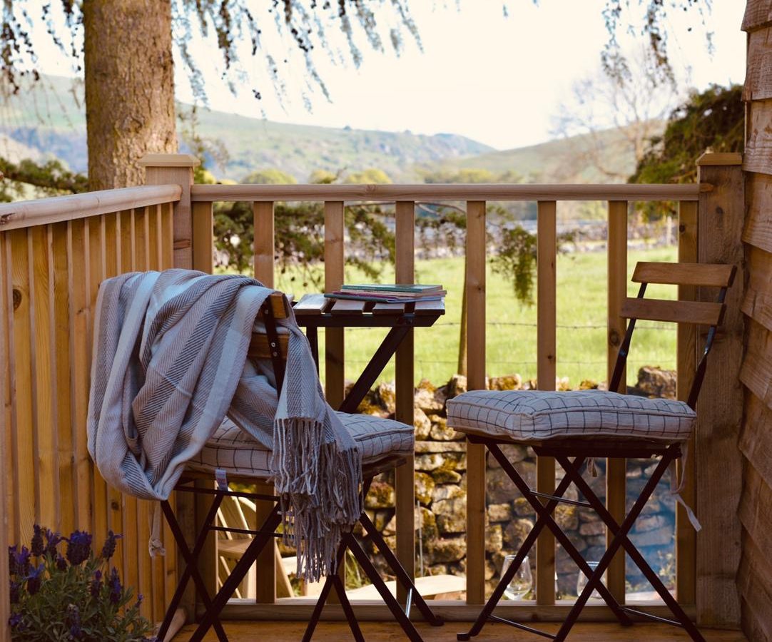 A cozy wooden deck at Smithyfields features two folding chairs with cushions, a small table with books, and a striped blanket draped over one chair. A lush, green landscape with hills is visible in the background. By the railing sits a potted lavender plant, adding a touch of color reminiscent of Shepherds Huts.