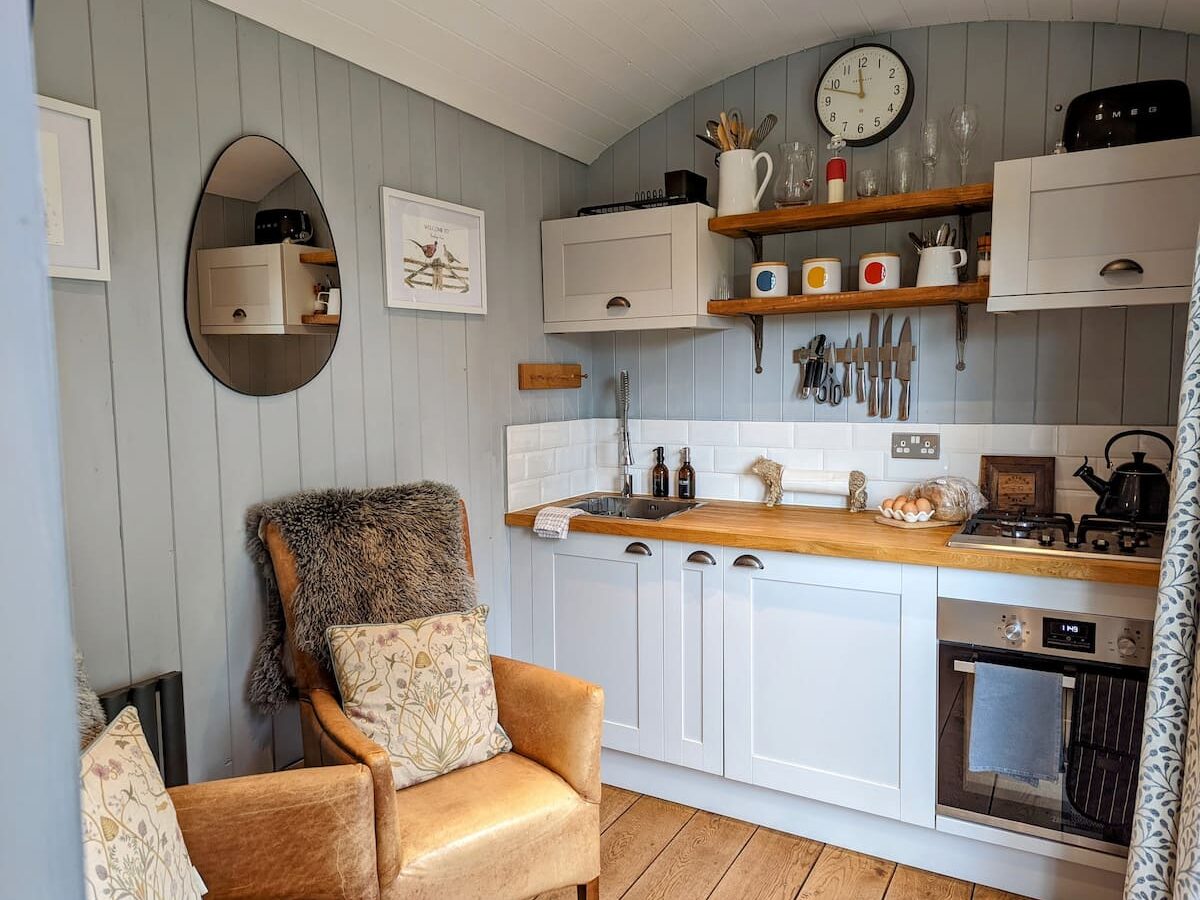 Cozy kitchen reminiscent of a Shepherds Hut, featuring wooden countertops, white cabinets, wall shelves adorned with a clock, and a plush brown armchair draped with a fur throw and cushions. The rustic charm offers an inviting retreat akin to the warmth of Smithyfields.