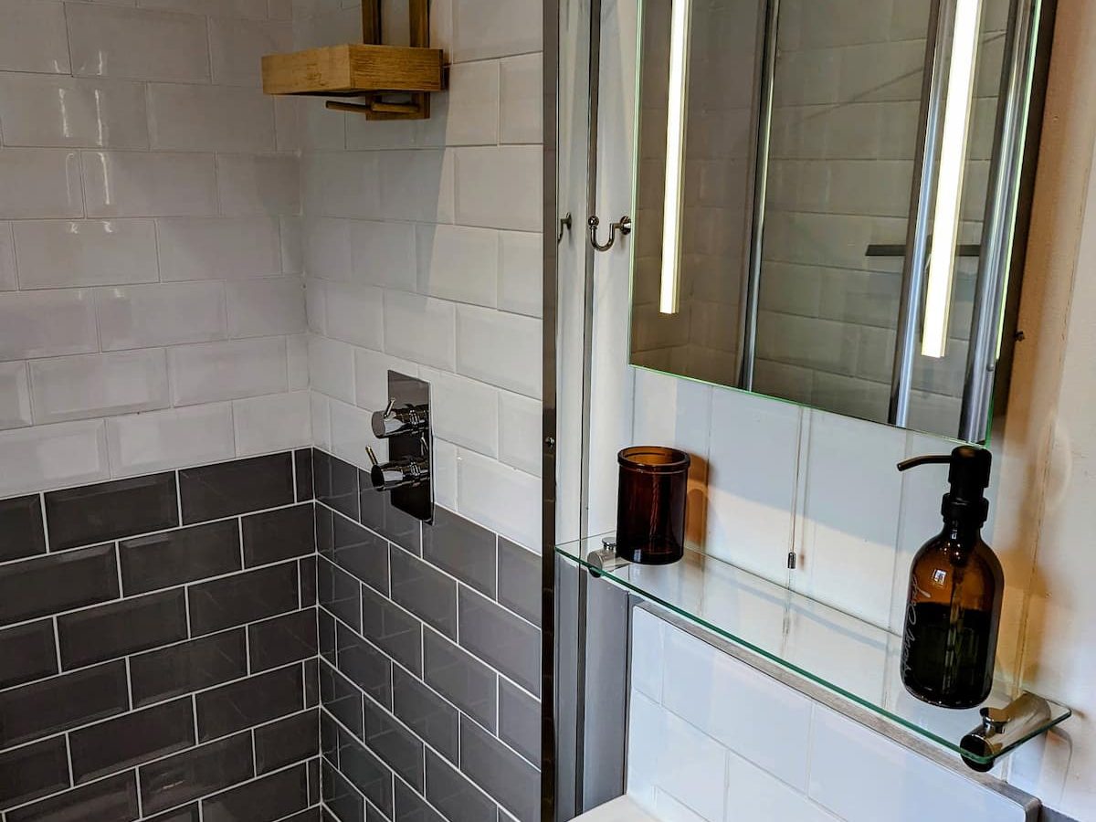 A modern bathroom featuring a small white sink with a chrome faucet and a mirrored medicine cabinet with integrated lighting above. Between the sink and cabinet, a small glass shelf holds a brown bottle. The shower, inspired by Smithyfields Shepherds' aesthetic, boasts white and dark gray subway tiles and a wooden shelf.