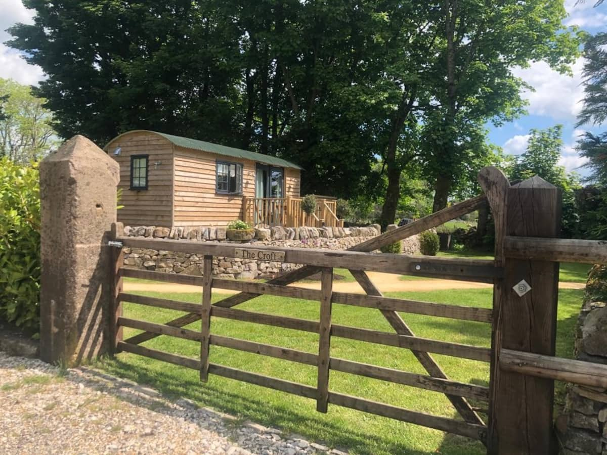 A charming wooden shepherd's hut is situated in a lush green yard, surrounded by large trees. The hut, part of the Smithyfields collection, resides behind an open wooden gate attached to a stone wall, with a sign reading 