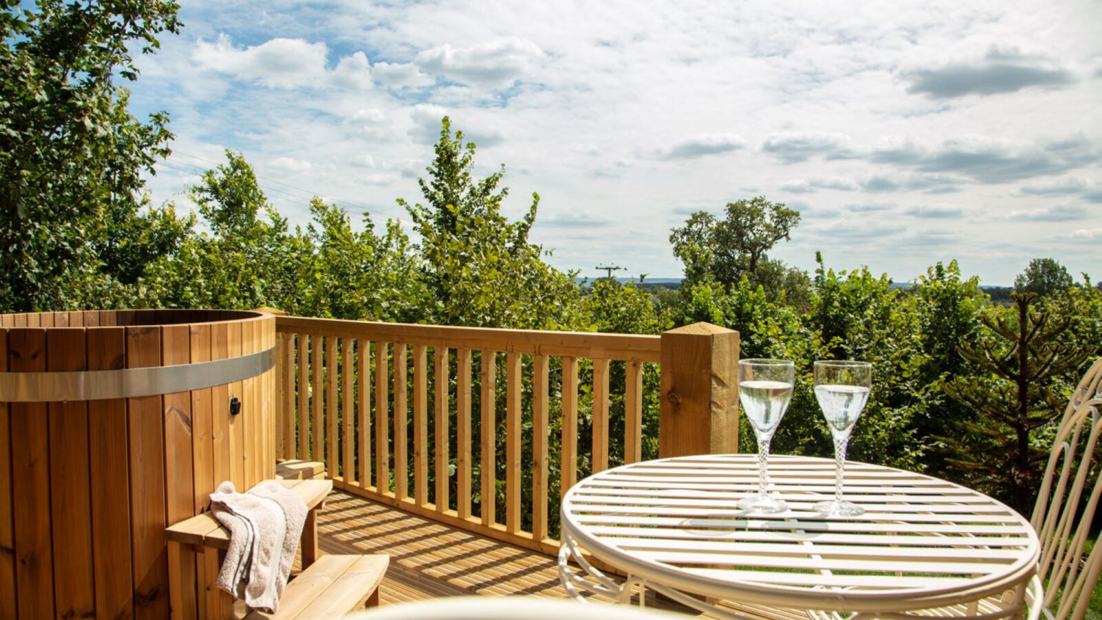 Sunny deck with a wooden hot tub and a white metal table set alongside two chairs, perfect for savoring glasses of water. The idyllic backdrop of trees evokes that serene, pastoral charm reminiscent of Abbey Farm's picturesque surroundings near the Shepherds Huts.