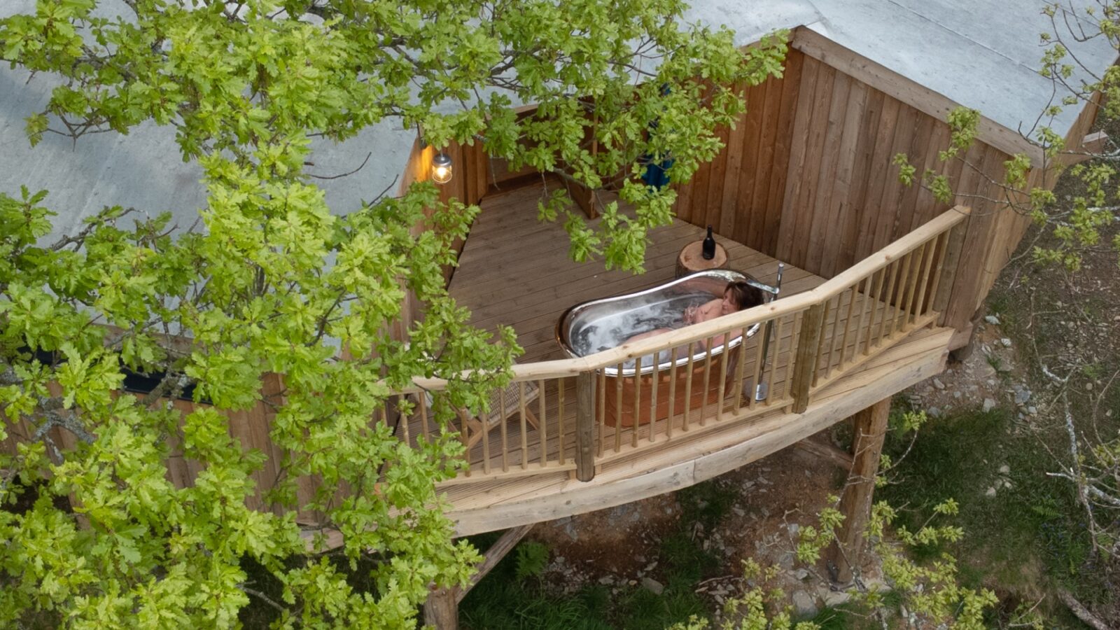 A person relaxes in a wild bathtub on a wooden balcony surrounded by lush greenery next to a rustic cabin, evoking the charm of Welsh treehouses.