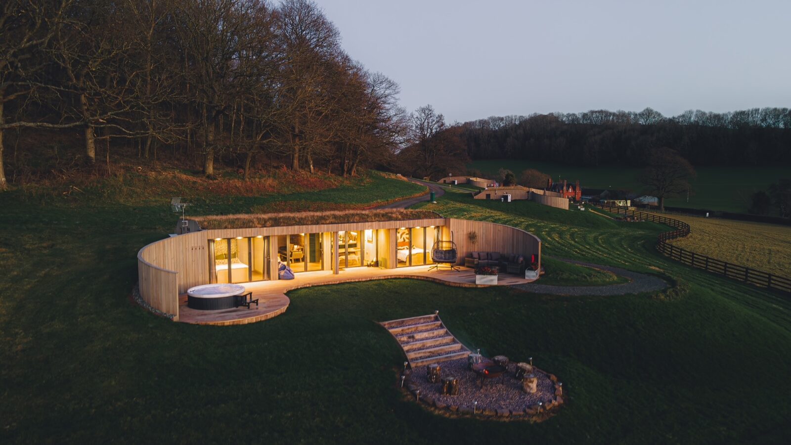 At dusk, the Pontypinna Hideaways reveal a modern, circular house with large windows, beautifully illuminated and nestled among grassy hills and trees.