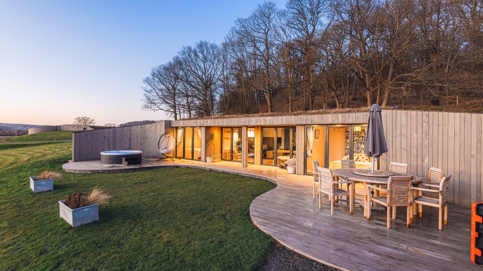 Discover the Pontypinna Hideaways: modern houses with large windows, a wooden deck, an outdoor dining set, potted plants, and stunning views of bare trees at sunset.