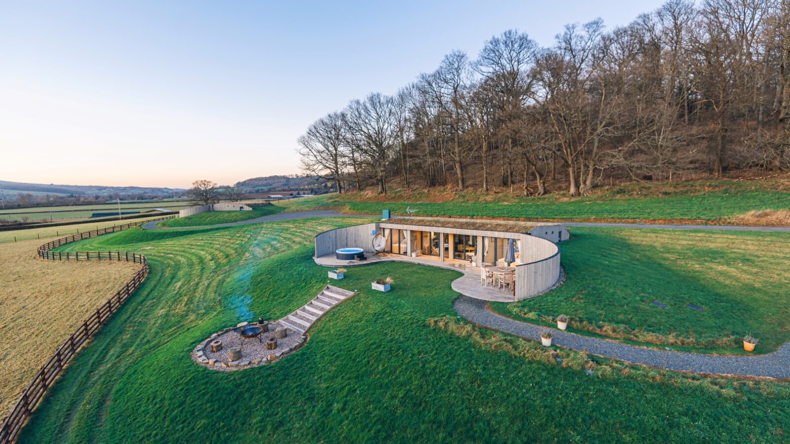 Nestled in the serene landscape of Pontypinna Hideaways, this modern circular house boasts a grass roof, seamlessly blending with the lush surroundings and sparse trees beneath a clear sky.