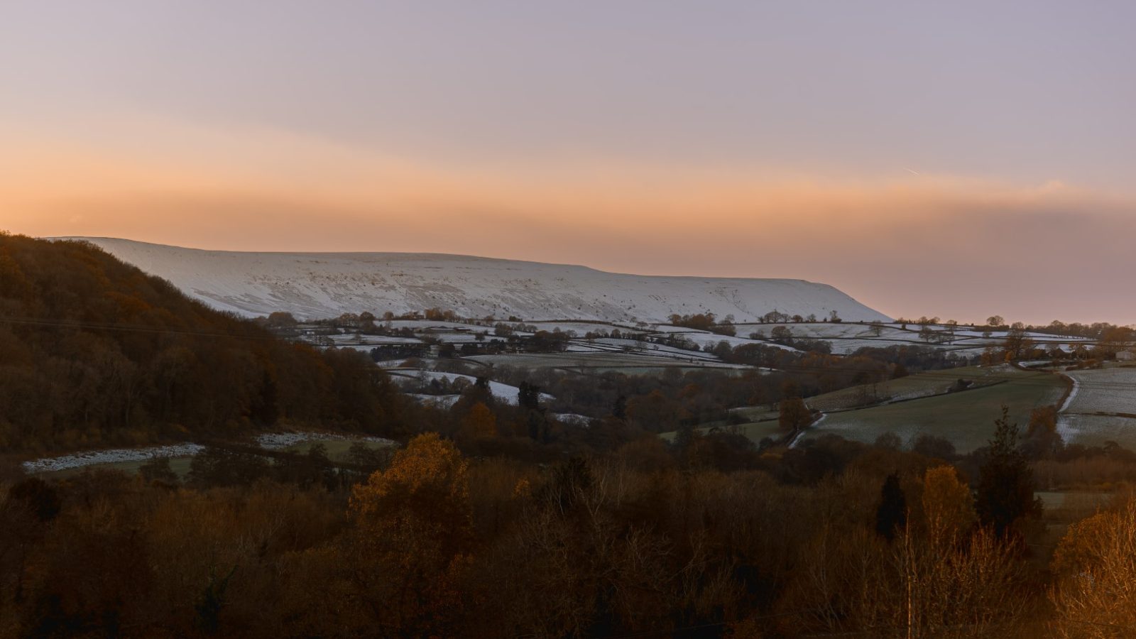 Snow-covered hill and fields under a pastel sky at sunrise, near Pontypinna Hideaways, surrounded by trees and countryside.