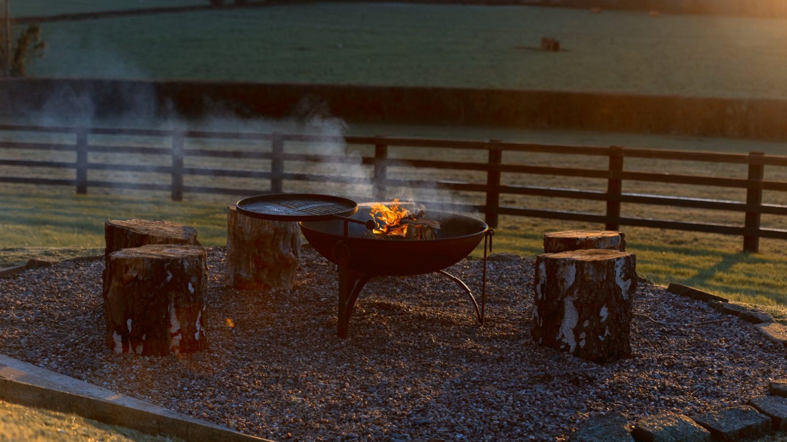 A fire pit with a grill is surrounded by log seats on gravel at Pontypinna Hideaways, set against fields and a wooden fence.