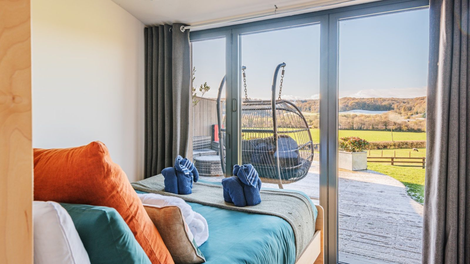 Pontypinna Hideaways' bedroom with a large window, towel origami on the bed, and patio view with hanging chairs over fields.