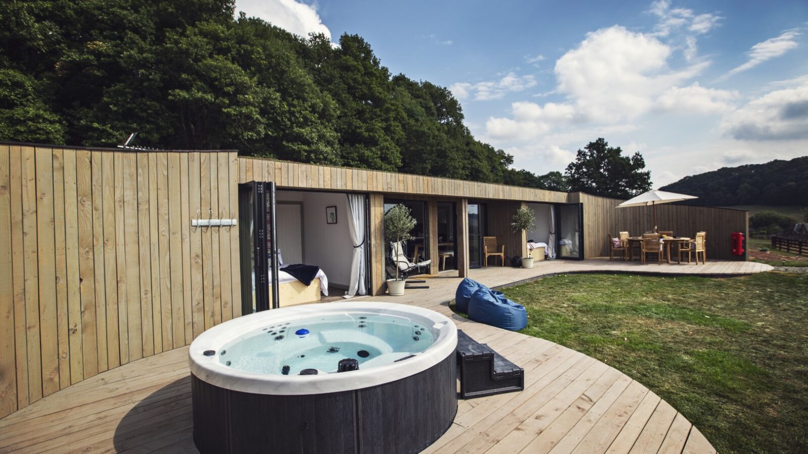 Deck with a hot tub in the foreground, surrounded by rustic Pontypinna wooden structures and trees, offering an idyllic hideaway. Cozy outdoor seating sits under a partly cloudy sky, perfect for relaxation and unwinding.