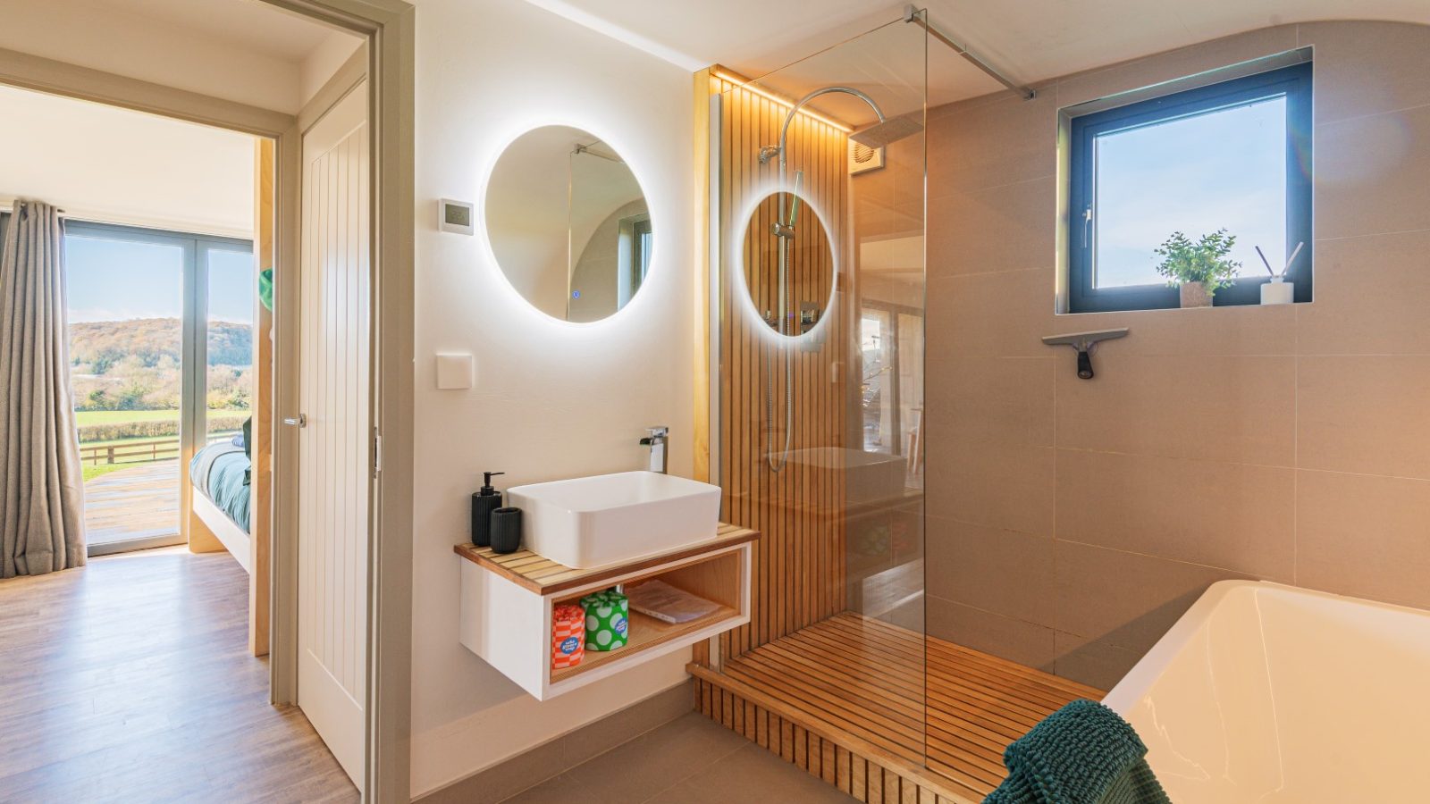 Modern bathroom at Pontypinna Hideaways features a round mirror, illuminated shower, and wooden accents. Bedroom with large windows seen through the door.