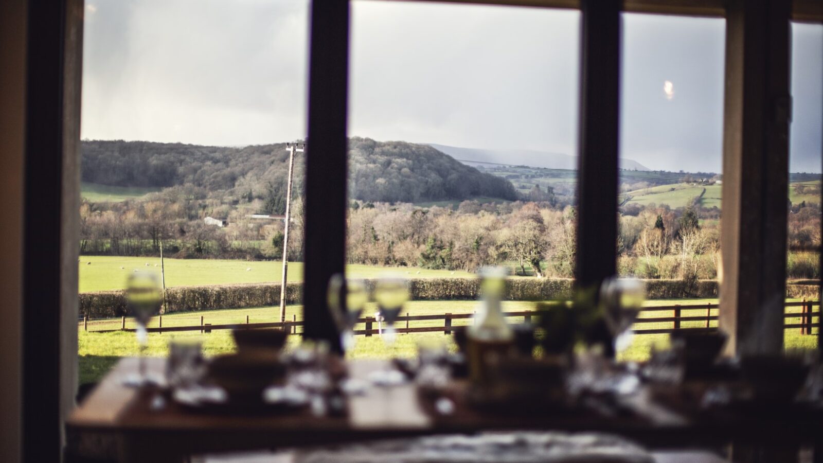 A scenic countryside view through large windows captures the essence of tranquil hideaways, with an out-of-focus table set for a meal in the foreground, evoking the charm of Pontypinna.