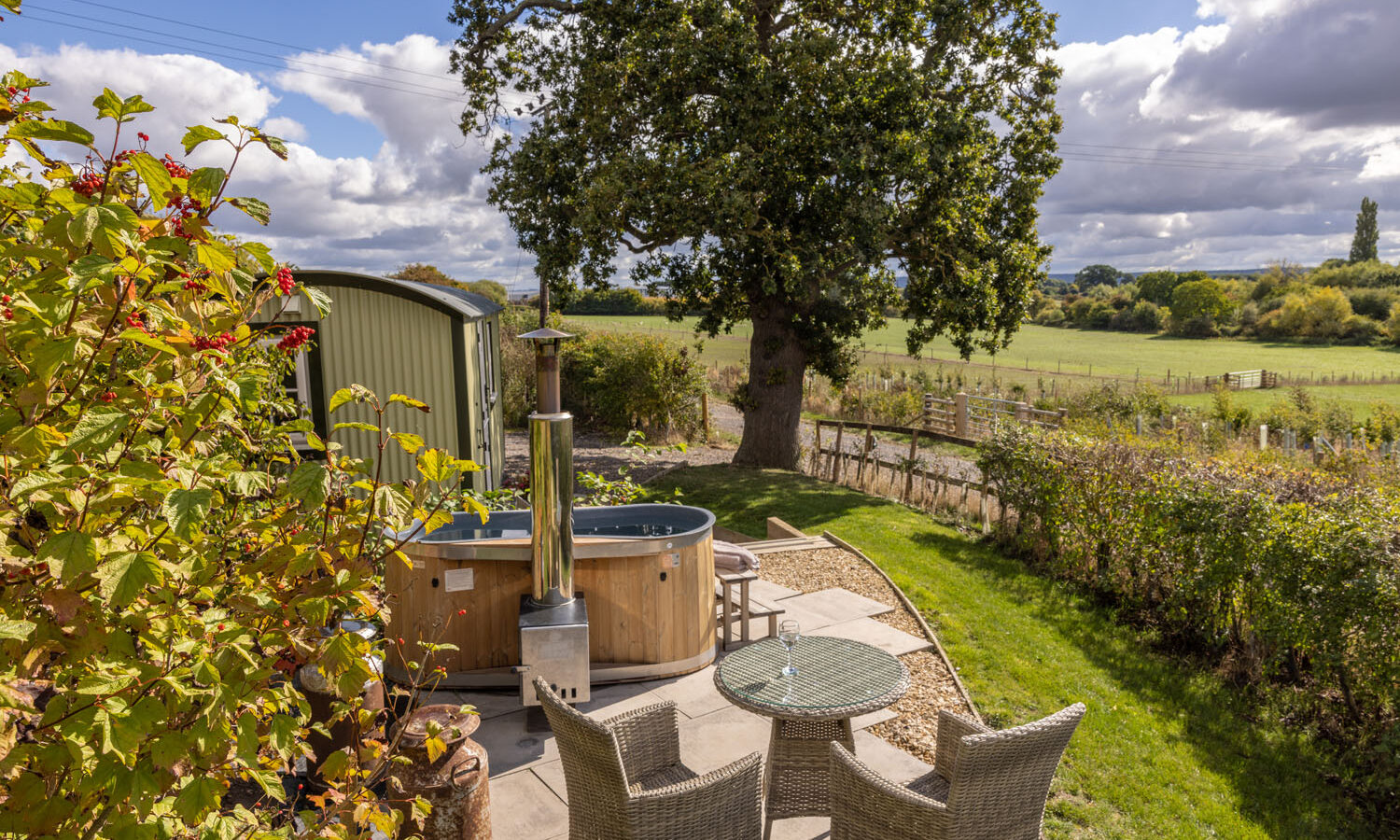 Outdoor seating with wicker chairs and table next to a wooden hot tub, overlooking the grassy fields of Abbey Farm and charming Shepherds Huts under a cloudy sky.