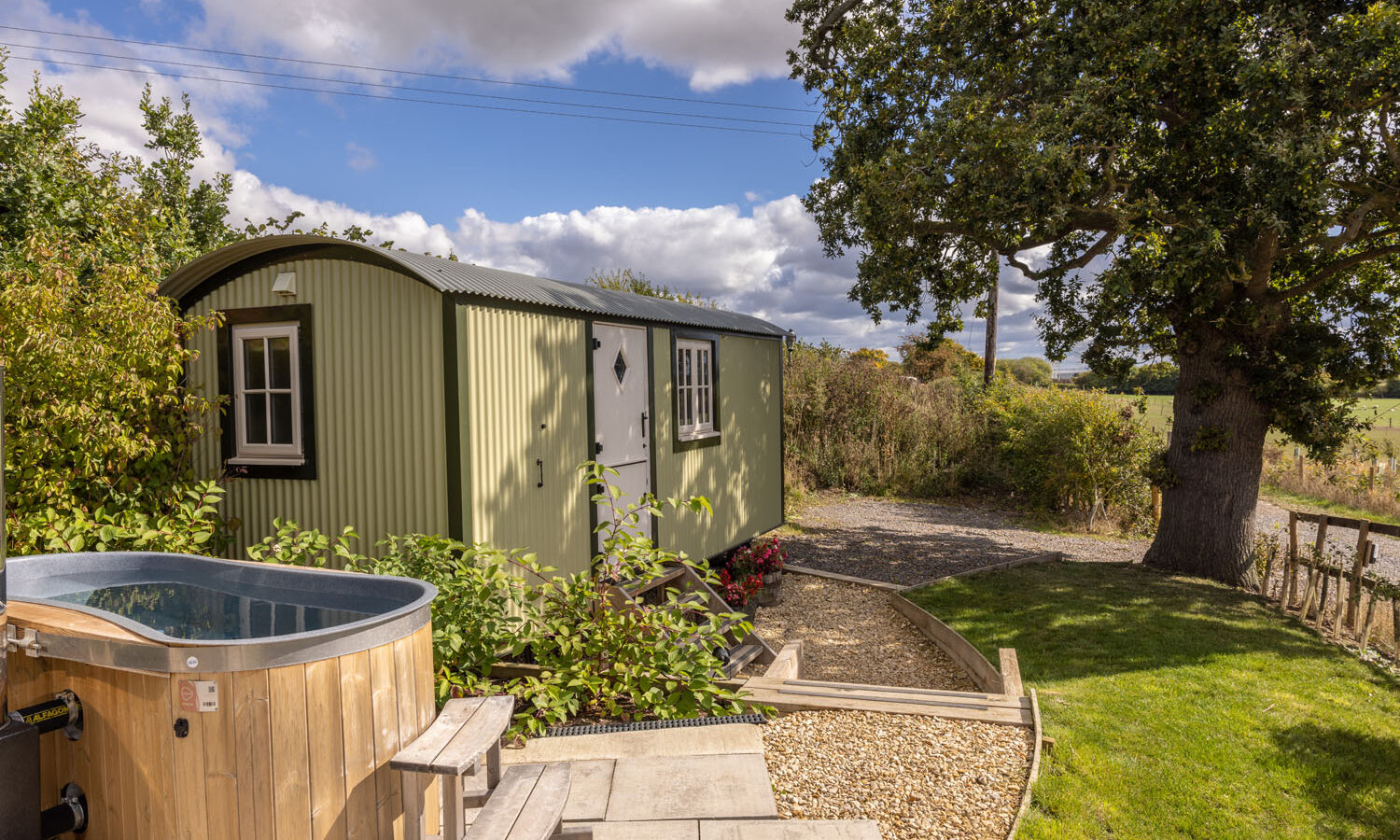 Nestled amidst verdant surroundings and towering trees under a cloudy sky, this rustic green cabin at Abbey Farm features a hot tub on a wooden deck, reminiscent of charming shepherds huts.