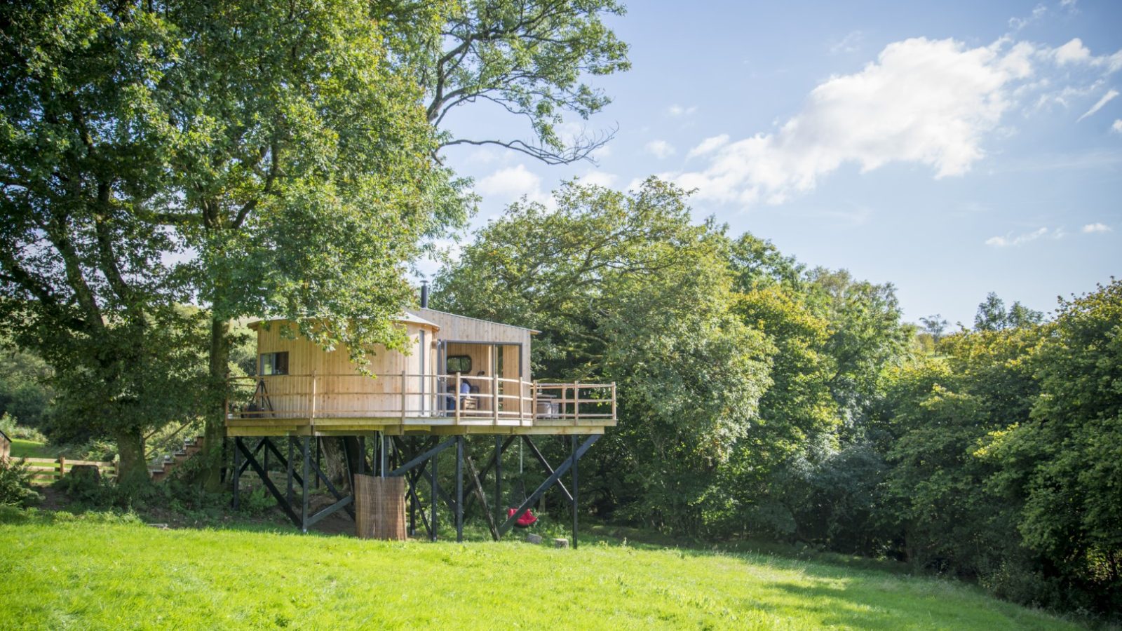 A small wooden treehouse elevated on stilts, nestled among lush green trees in a wild, Welsh landscape. It features a wraparound balcony with views of the grassy field under a clear blue sky. A red hammock swings gently beneath, inviting relaxation amid nature's embrace.
