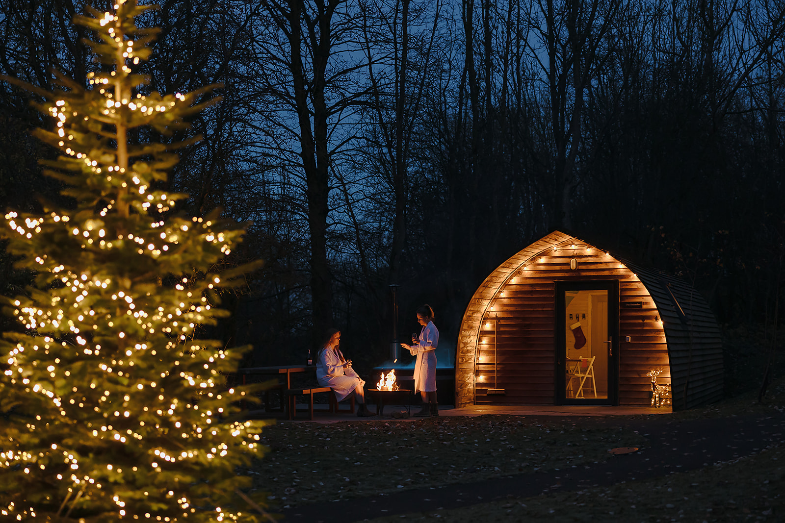 A cozy cabin at night with two people by a fire pit offers one of the coolest Xmas getaways in the UK. A decorated tree with lights twinkles in the foreground, creating a magical holiday atmosphere.